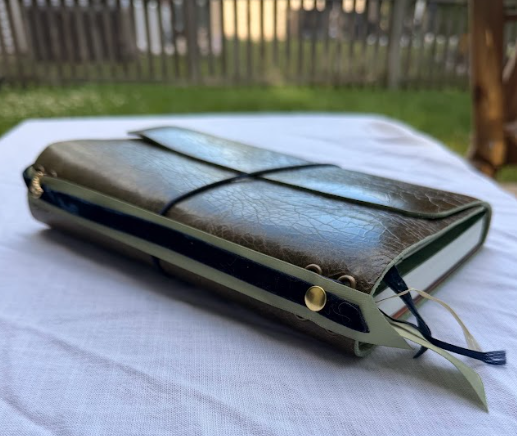 closed leather-bound journal with a strap, a pen and a ribbon marker on a white cloth surface outdoors.