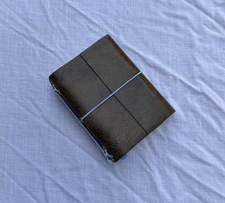 Black leather wallet with light blue elastic band on a light purple fabric surface.