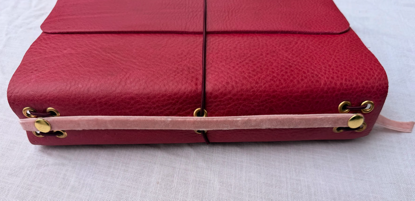 Pink leather journal with gold eyelet accents and pink strap detail on a white surface.
