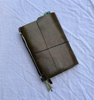Green leather notebook or journal with elastic band, green ribbon bookmark, and zipper pocket, placed on a light-colored fabric background.
