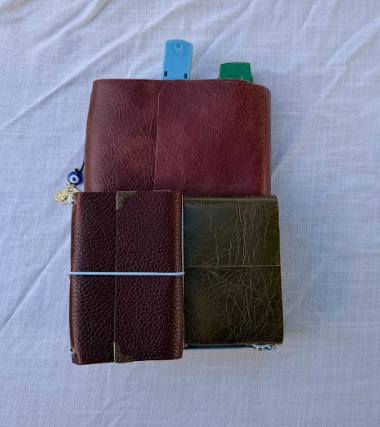 Three notebooks stacked on a white fabric surface, with the top notebook in burgundy, the middle in dark green, and the bottom in purple accompanied by a blue and a green pen.