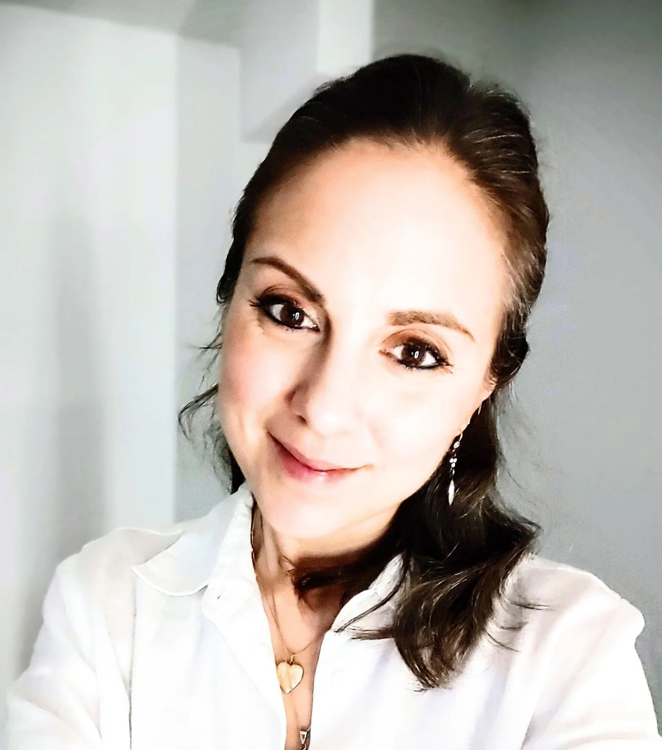Close-up of a smiling woman in a white shirt with dark brown hair styled in loose waves, wearing earrings and a heart-shaped pendant necklace, standing against a neutral background.