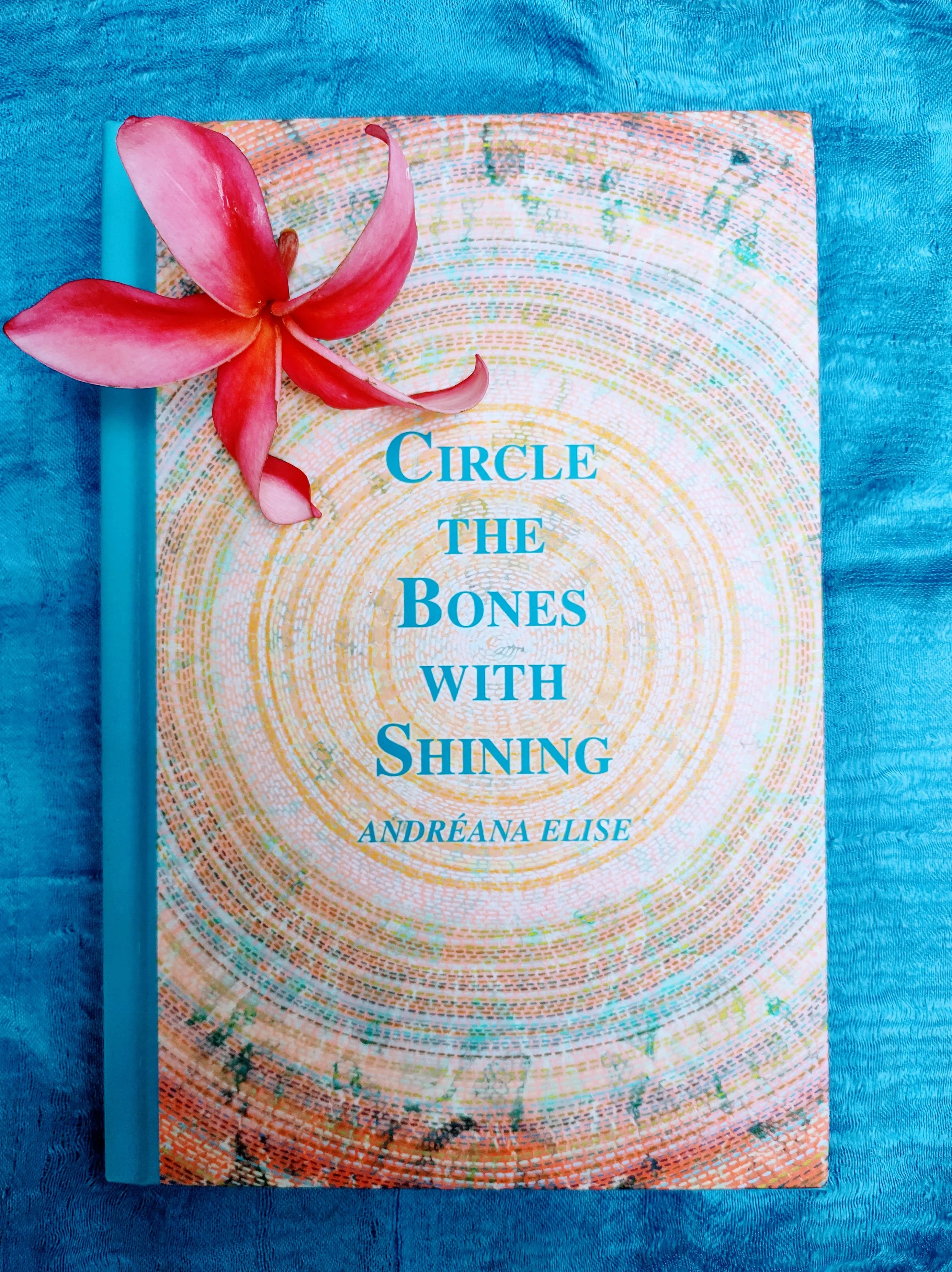 Book titled 'Circle the Bones with Shining' by Andreana Elise with a pink flower on top, placed on a blue textured surface.