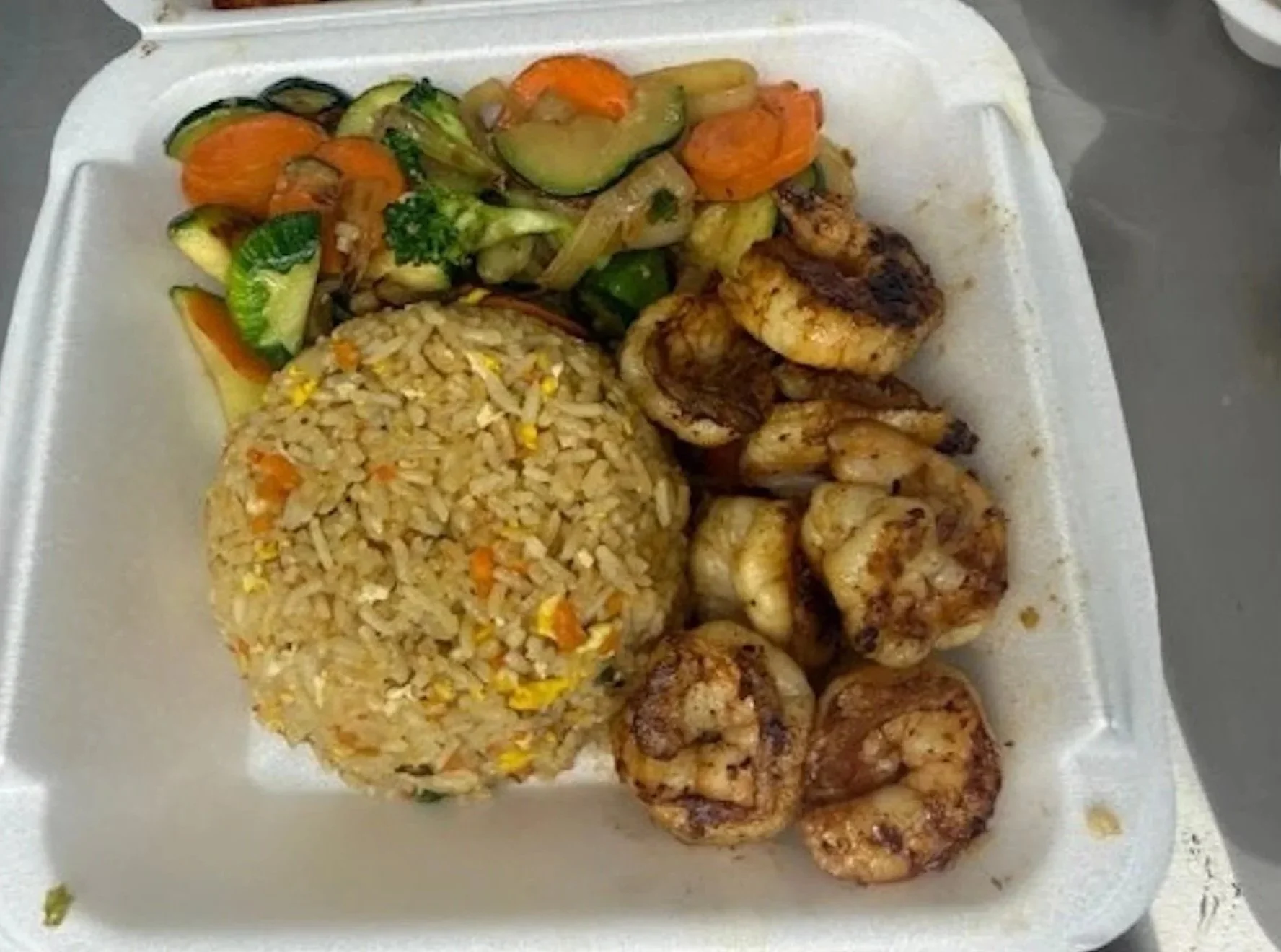 Takeout container with seasoned grilled shrimp, a mound of fried rice with vegetables, and a side of sautéed mixed vegetables including zucchini, carrots, and onions.