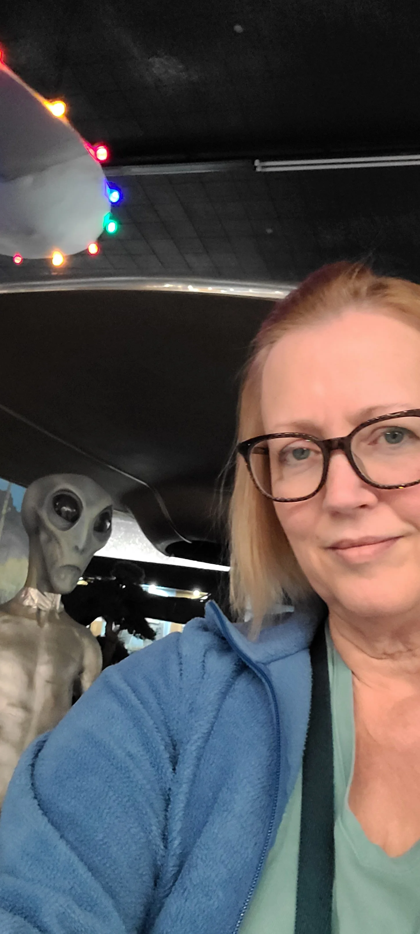 A woman with glasses taking a selfie next to a life-sized alien figure inside a location decorated with sci-fi themed decorations.