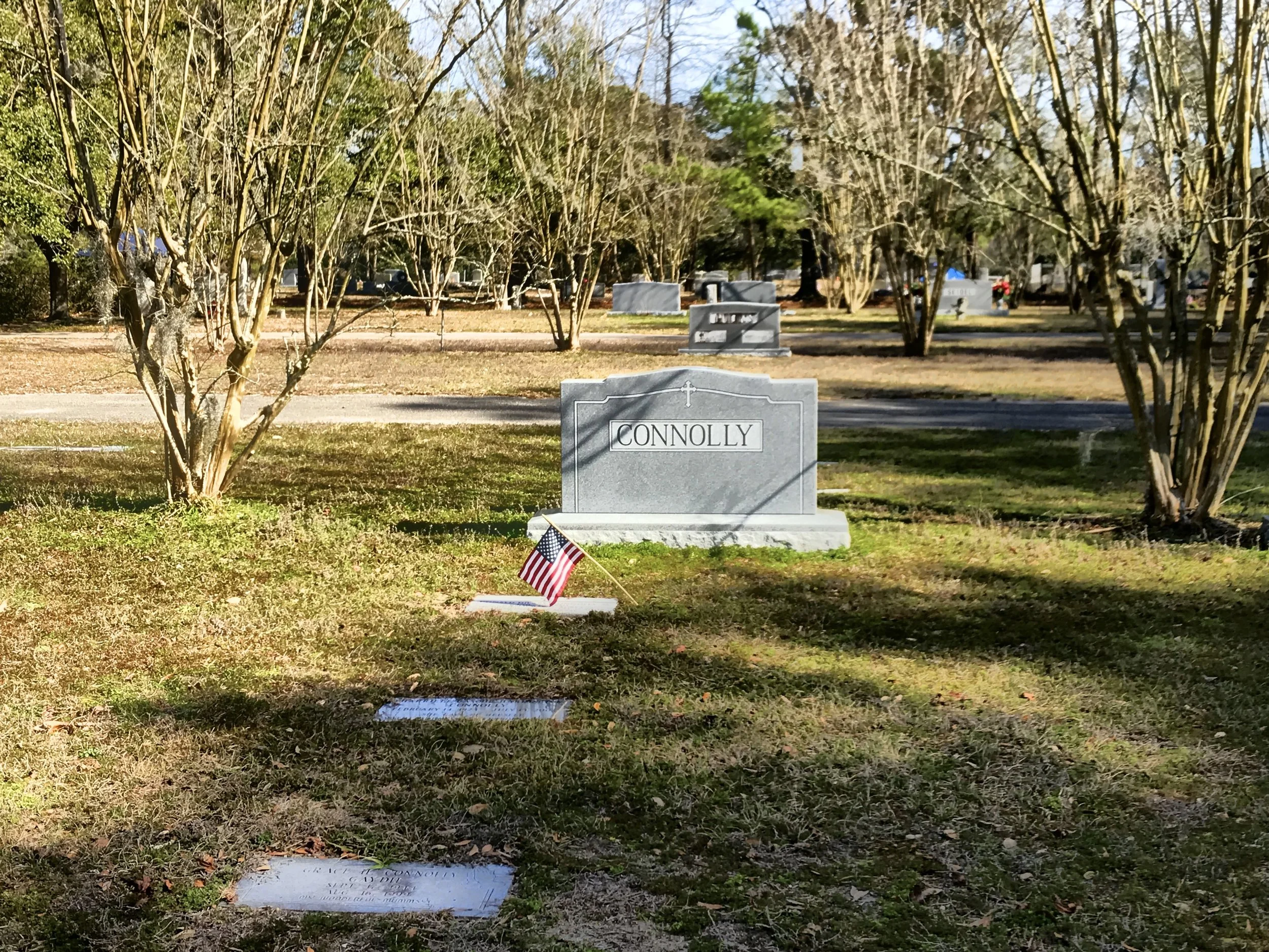 A cemetery scene with a gray headstone inscribed with the name 'CONNOLLY' amidst trees and grass, with a small American flag in front of the headstone and additional headstones visible in the background.