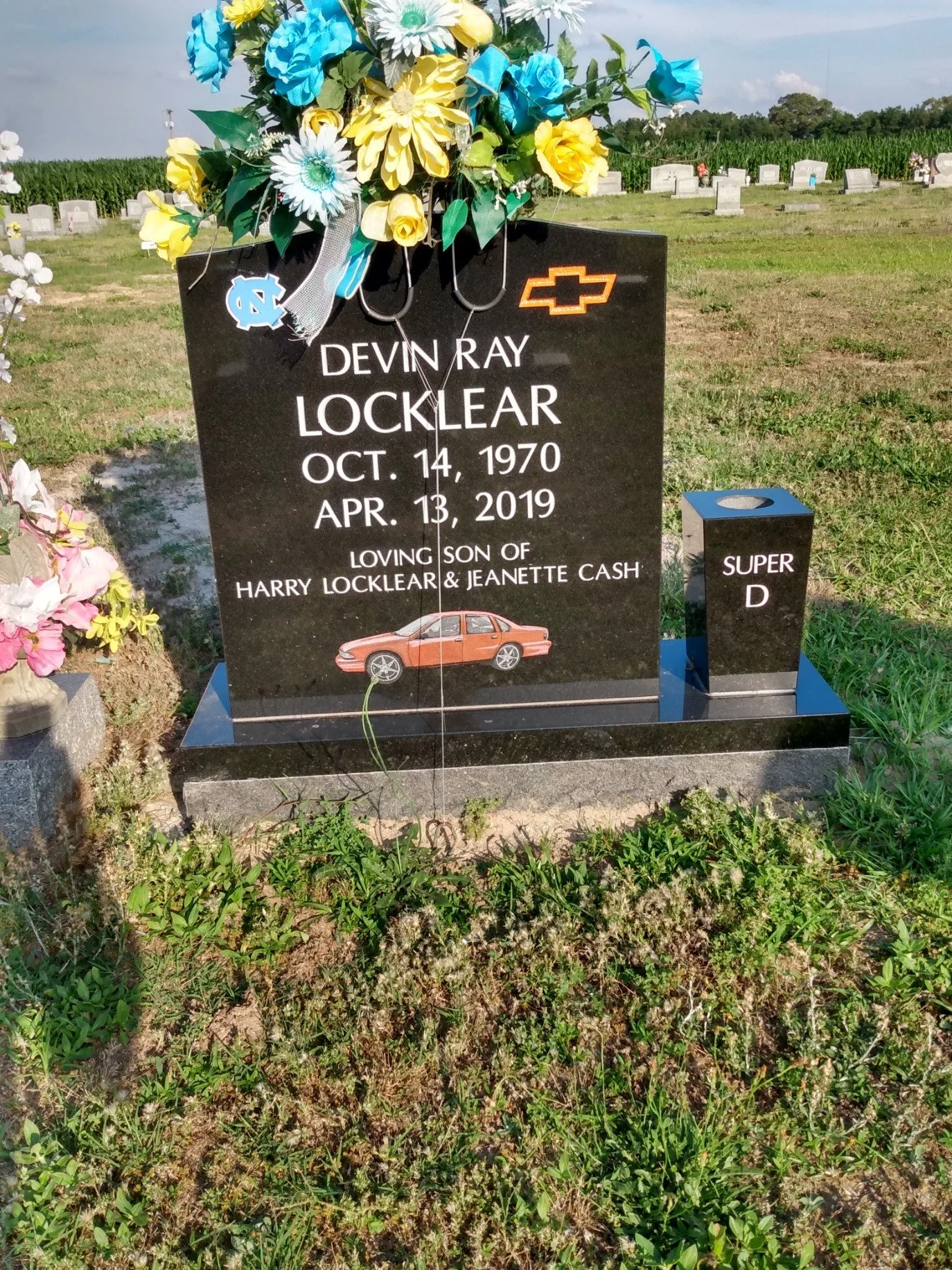 Gravestone with colorful artificial flowers and religious symbols, in a cemetery with other gravestones and a field of crops in the background.