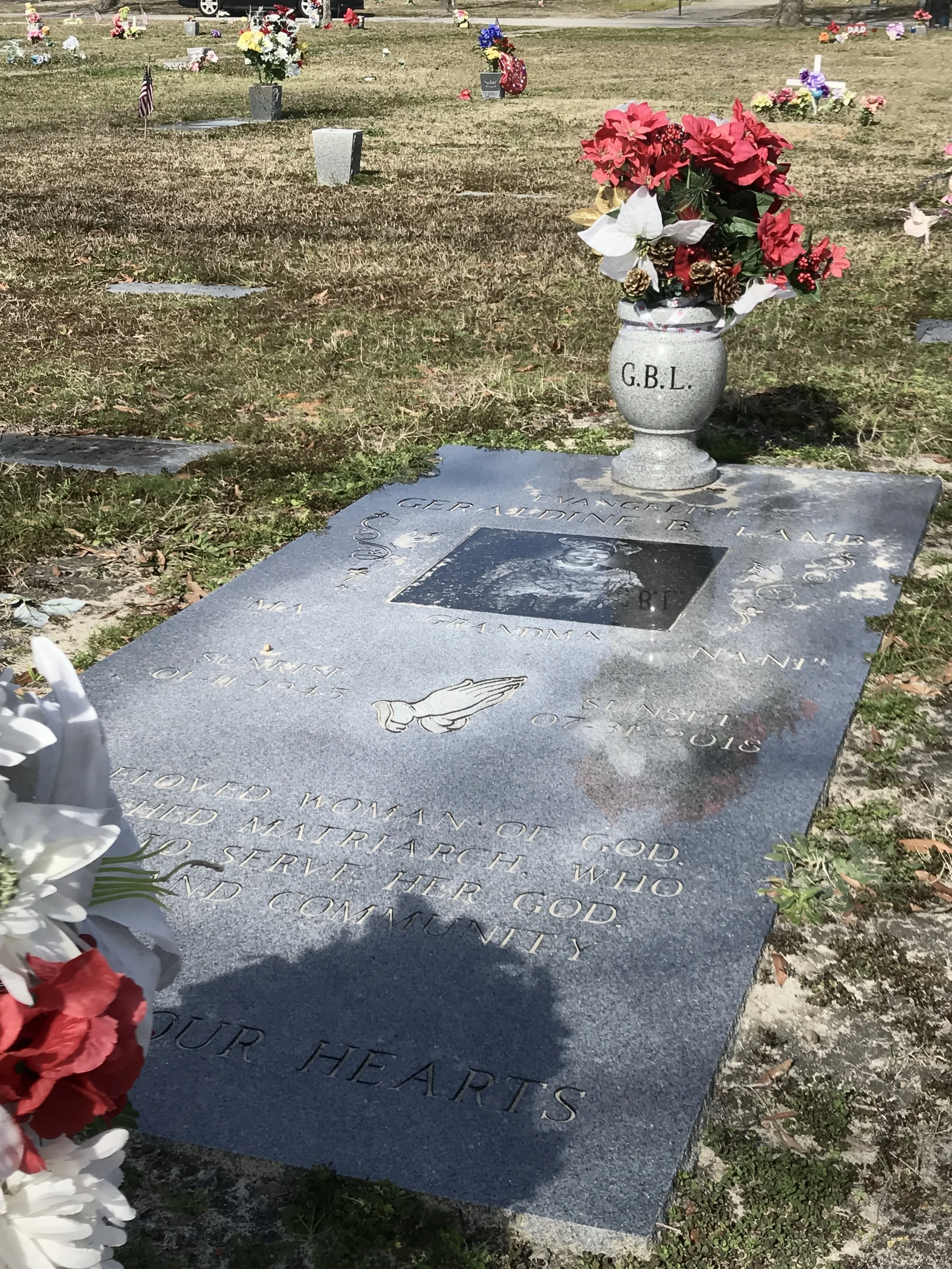 A gravestone in a cemetery with flowers and a small American flag. The gravestone inscribes the name Gertrude B. Hahn and a Bible verse. A large vase with red and white flowers and pinecones sits on the grave.