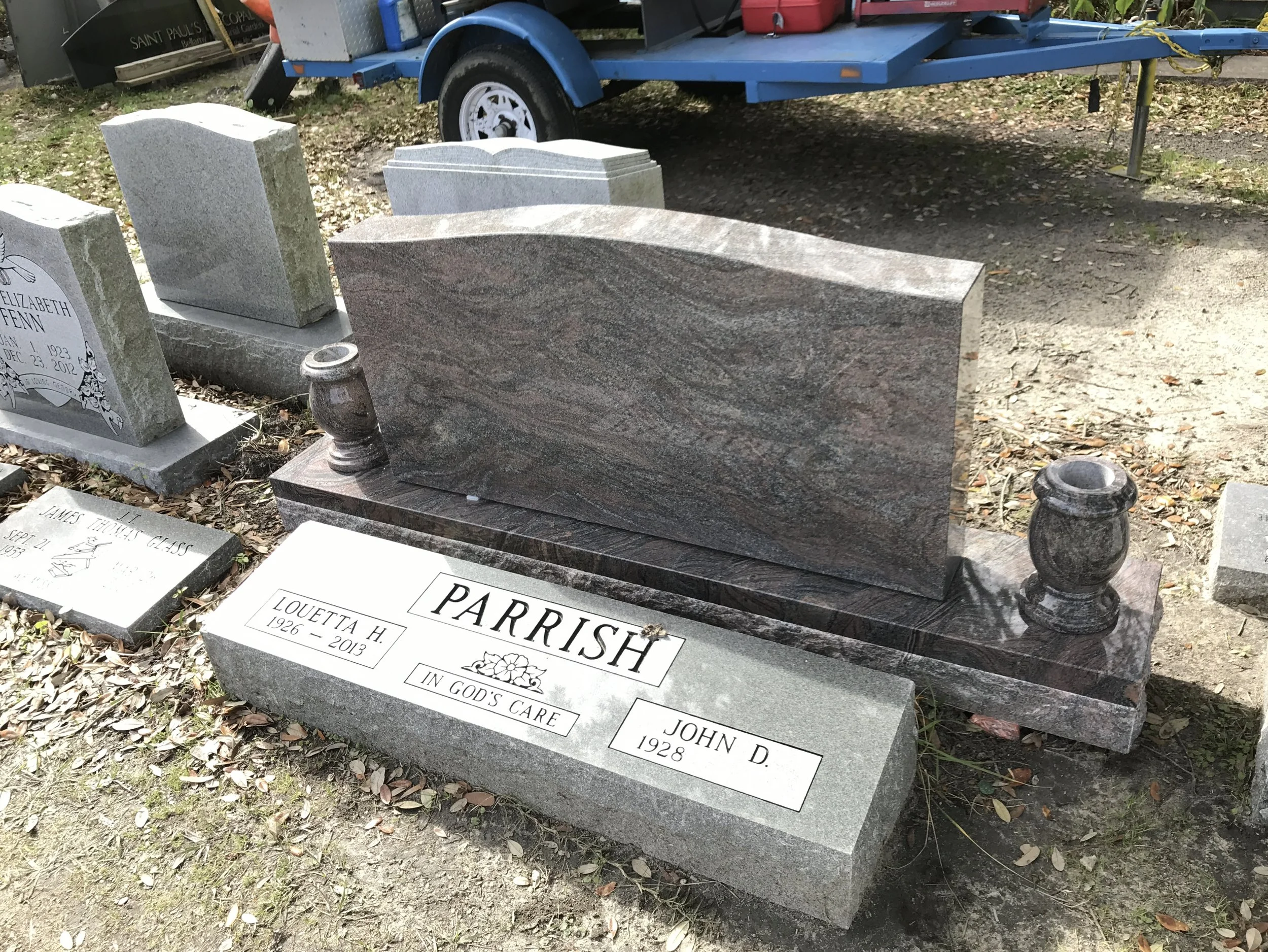 A granite headstone with the name Parrish in a cemetery, surrounded by other headstones. A blue trailer is in the background.