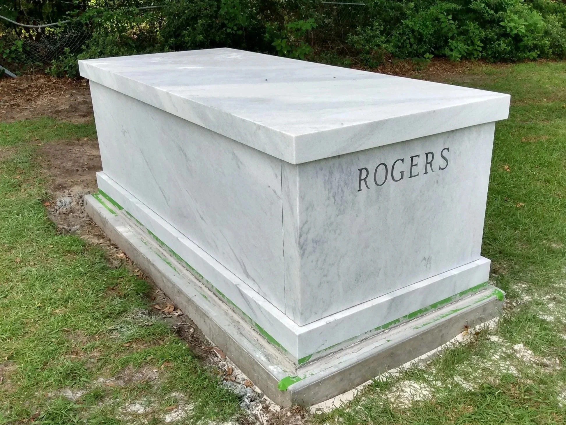 A white marble grave marker with the name 'ROGERS' engraved on it, located in a grassy area with some soil and green plants in the background.