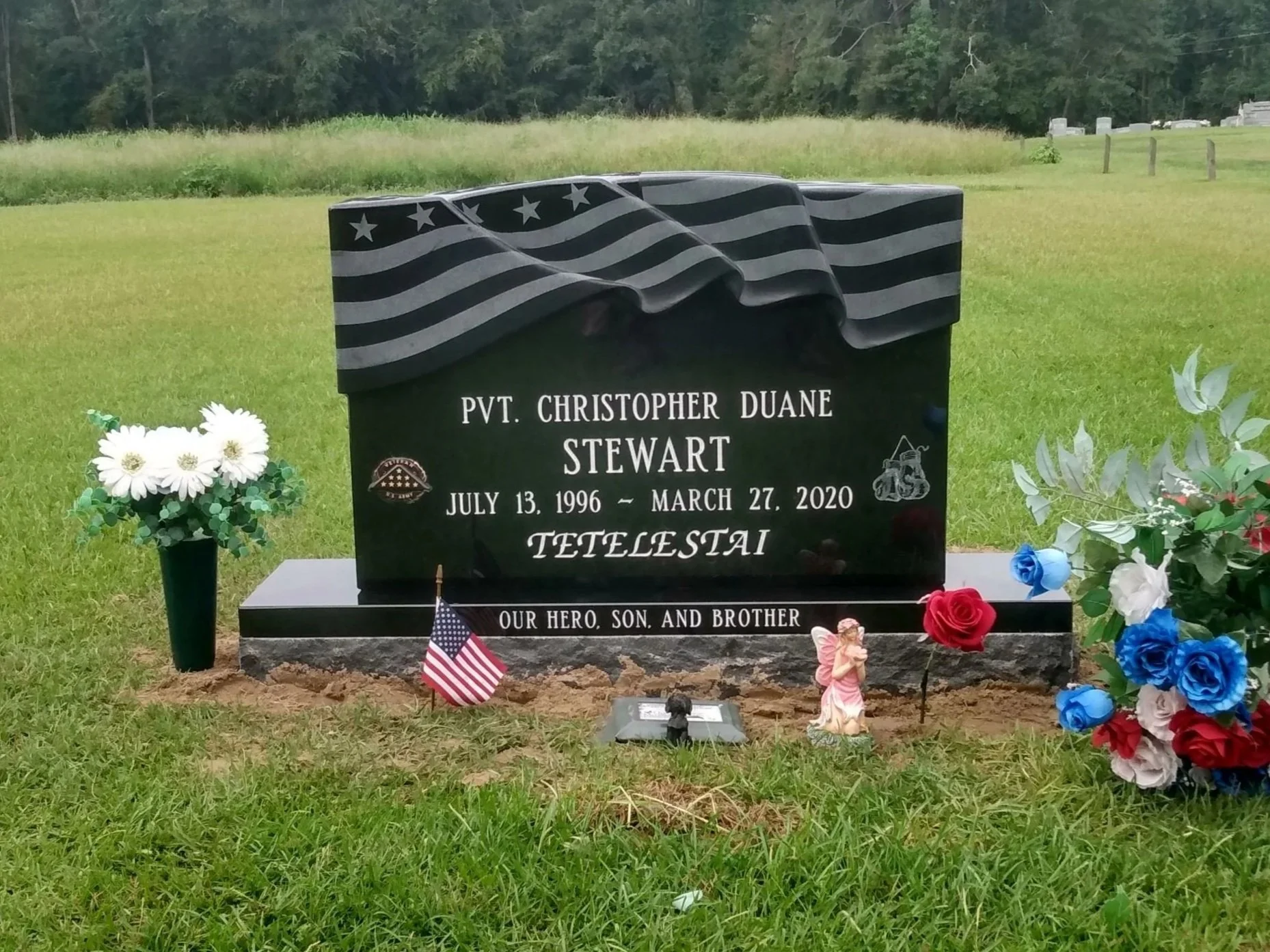 Gravestone with an American flag, flowers, and an angel figurine. The headstone honors Pvt. Christopher Duane Stewart (July 13, 1996 - March 27, 2020). It has a waving American flag engraved at the top and text reading, "Our hero, son, and brother."