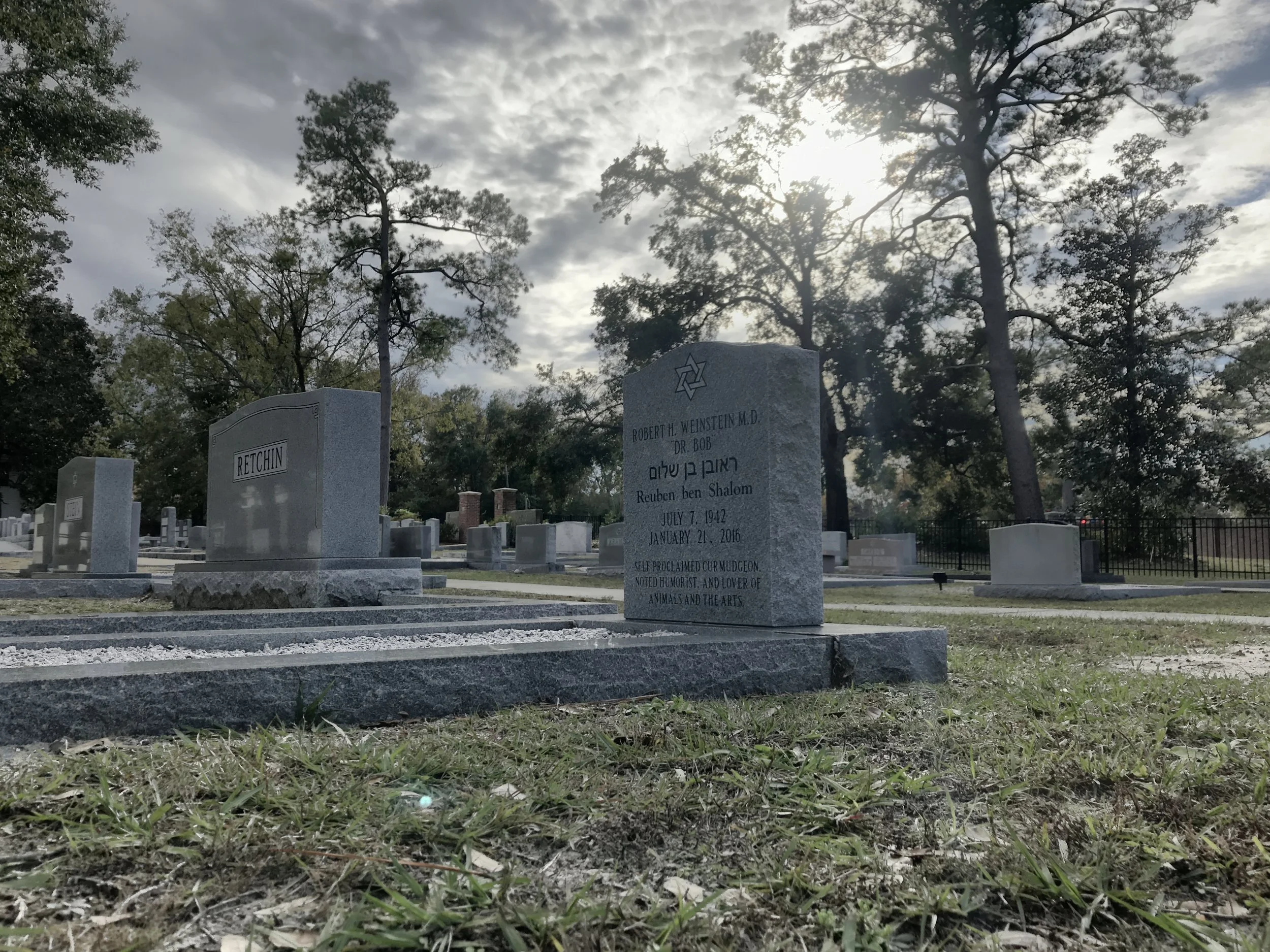 Gravestones in a cemetery with tall trees and cloudy sky in the background.
