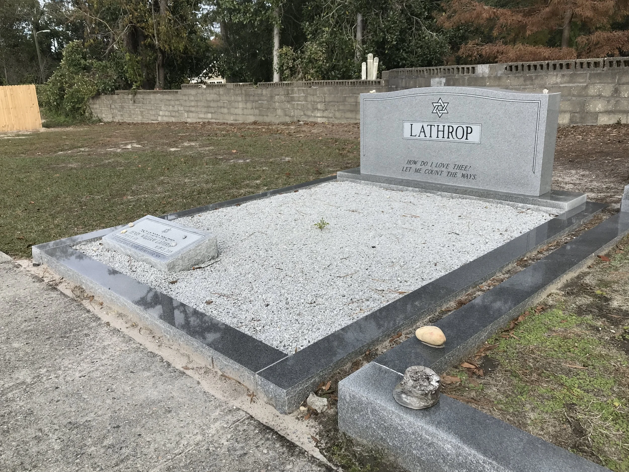A gravestone with the name "Lathrop" and a Star of David symbol, surrounded by gravel and a concrete border, in a cemetery with trees and a wooden fence in the background.