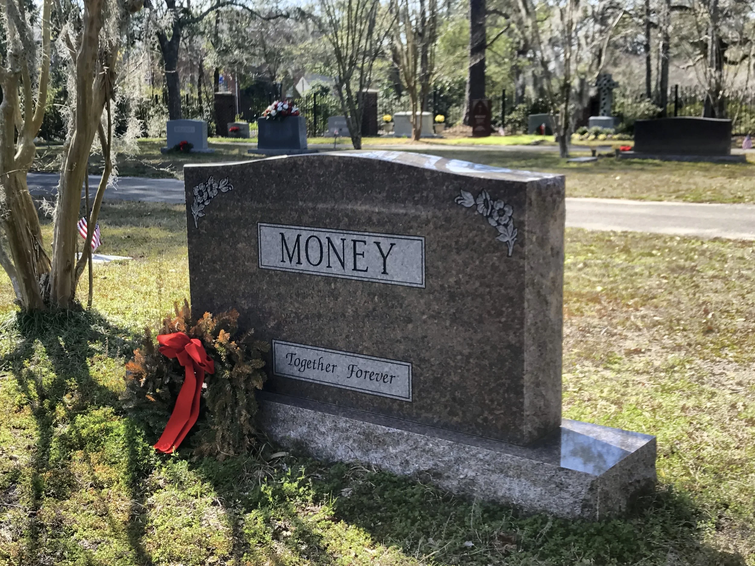 A granite gravestone with the word 'MONEY' engraved on it, decorated with engraved flowers on its corners, located in a cemetery with other headstones and memorials in the background, surrounded by trees and grass, with a small American flag and a re