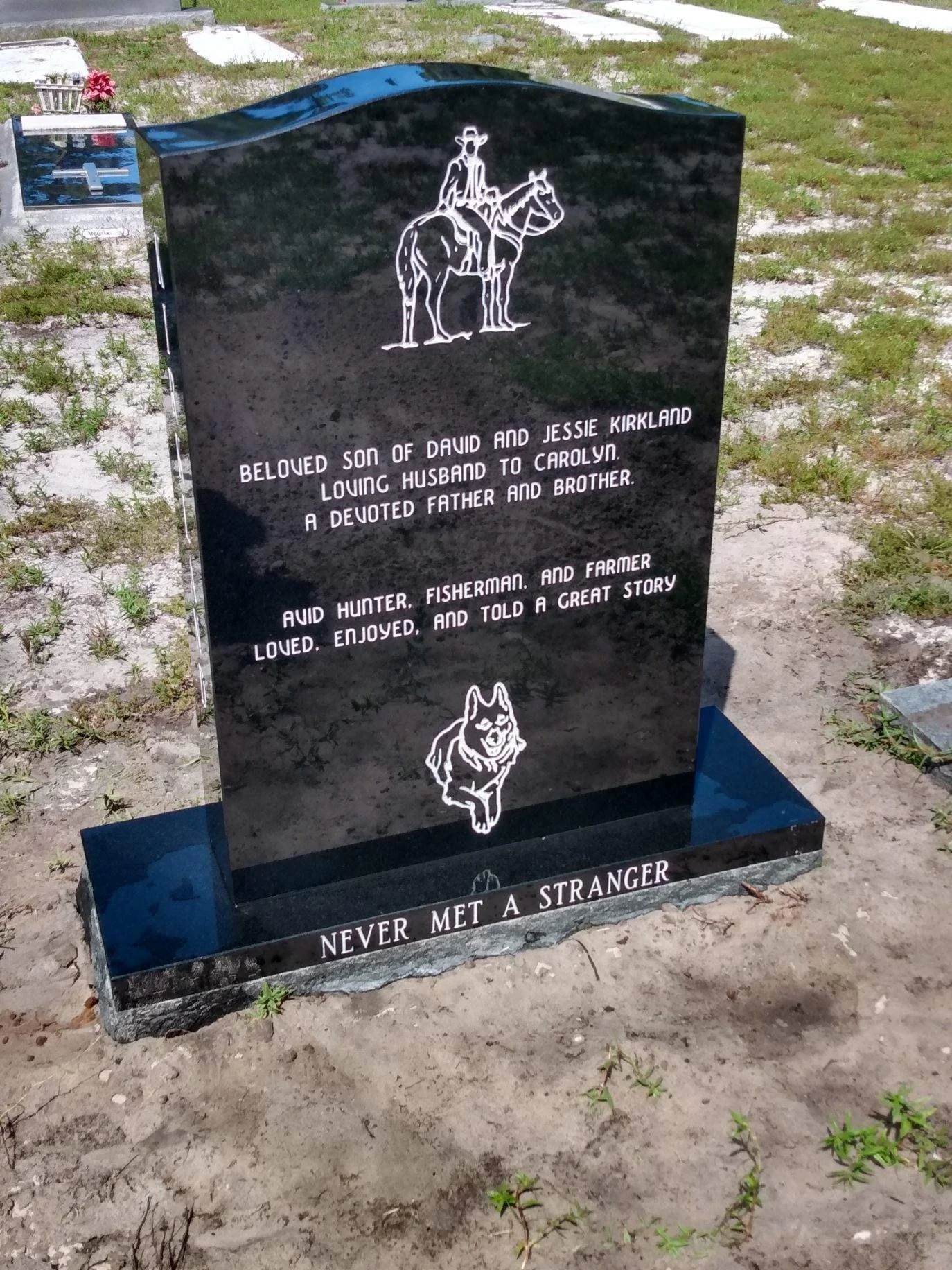Black granite headstone with a white engraving of a man riding a horse, wearing a hat. Text mentions a beloved son of David and Jessie Kirkland, devoted father and brother, and loved hunter, fisherman, and farmer. The bottom of the headstone reads: '