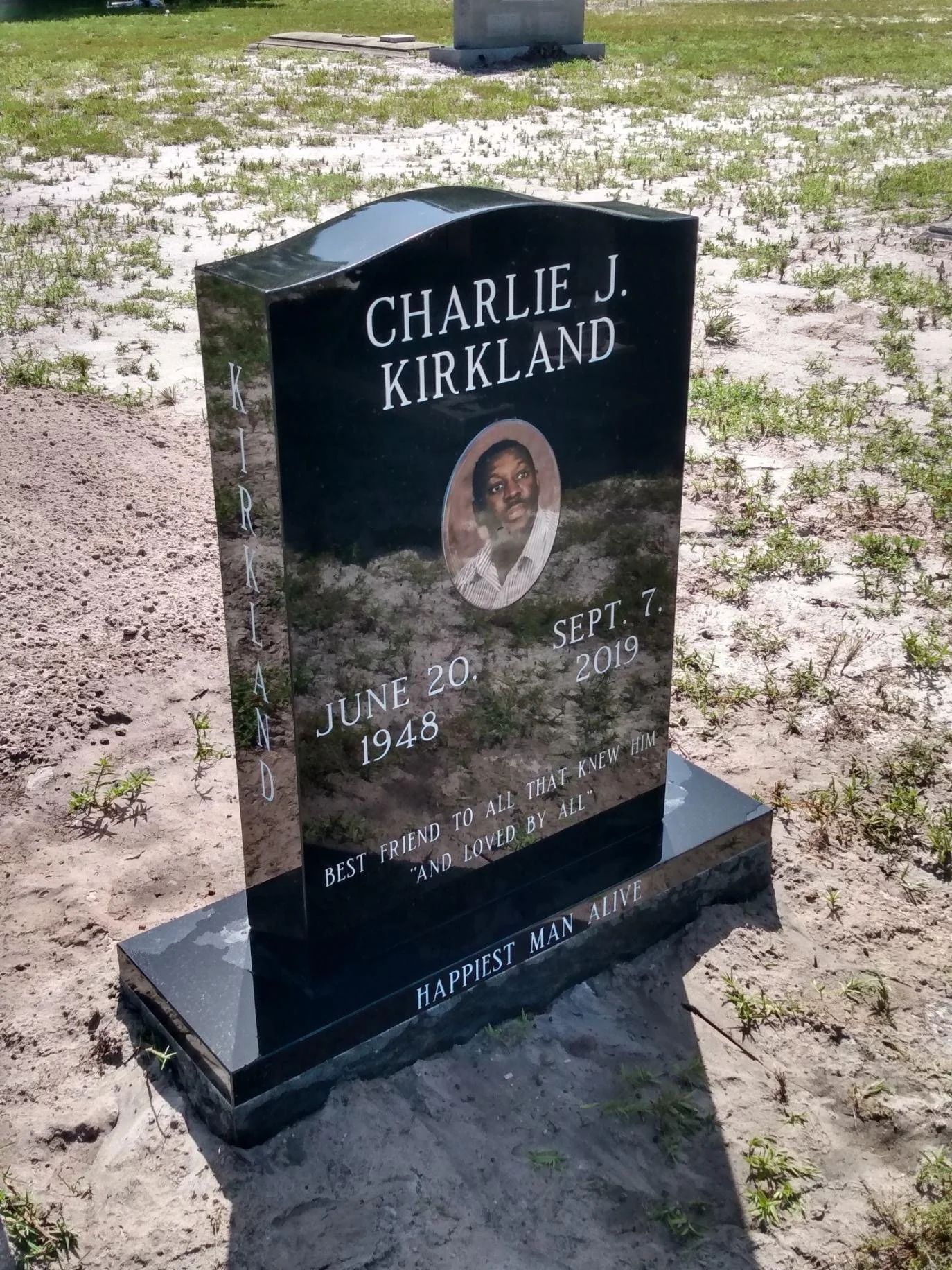 Black granite headstone with white lettering for Charlie J. Kirkland, including a color photo of him, birth date June 20, 1948, and death date September 7, 2019, on a patchy grassy area.