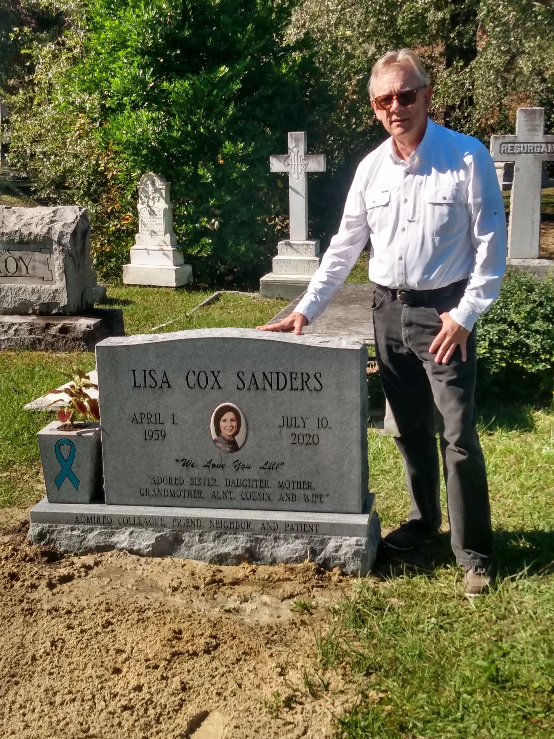 A man in sunglasses wearing a white shirt and black pants stands beside a gravestone with the name Lisa Cox Sanders. The gravestone features a photo, a ribbon, and inscriptions honoring her life and loved ones. The background includes additional grav