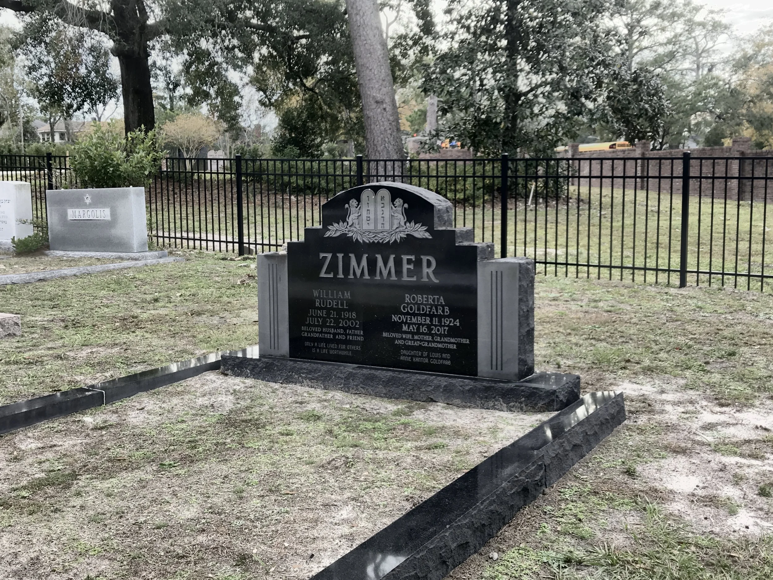 A cemetery with multiple gravestones, one of which has the name 'Zimmer' engraved, surrounded by a black metal fence, trees in the background, and a school bus visible through the trees.