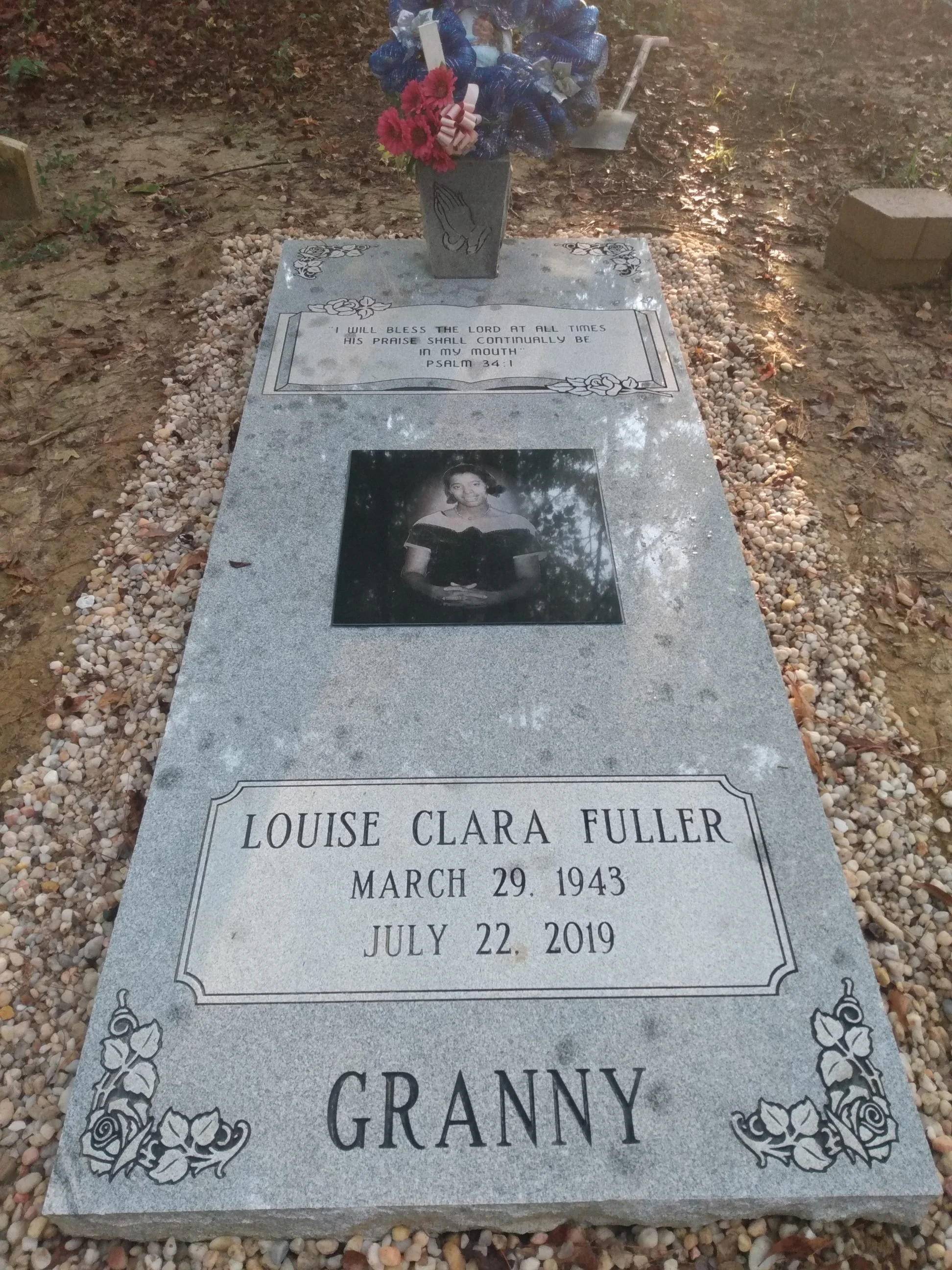 Gravestone with a small photograph of a young woman named Granny, born March 29, 1943, died July 22, 2019, surrounded by small pebbles. There are flowers and a photo of a small figurine on top of the gravestone.