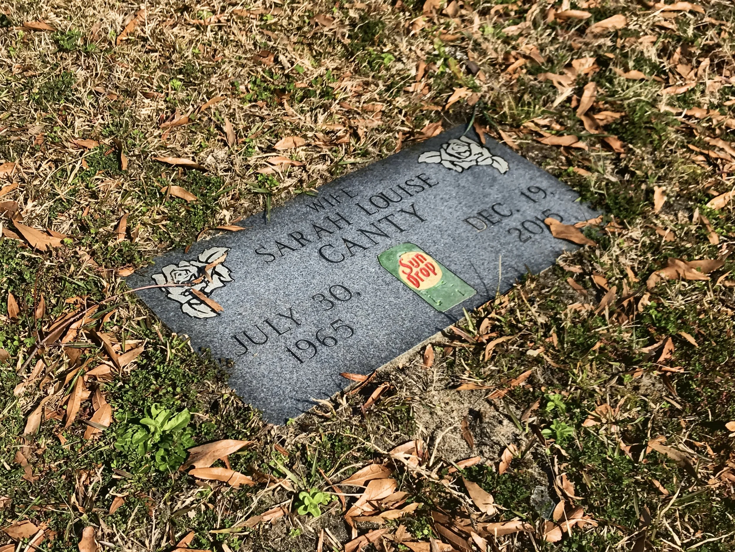 Gravestone for Sarah Louise Gill, who passed away on July 30, 1965, with a decorative rose and a small Sun Drop soda sticker on it, surrounded by grass and dried leaves.