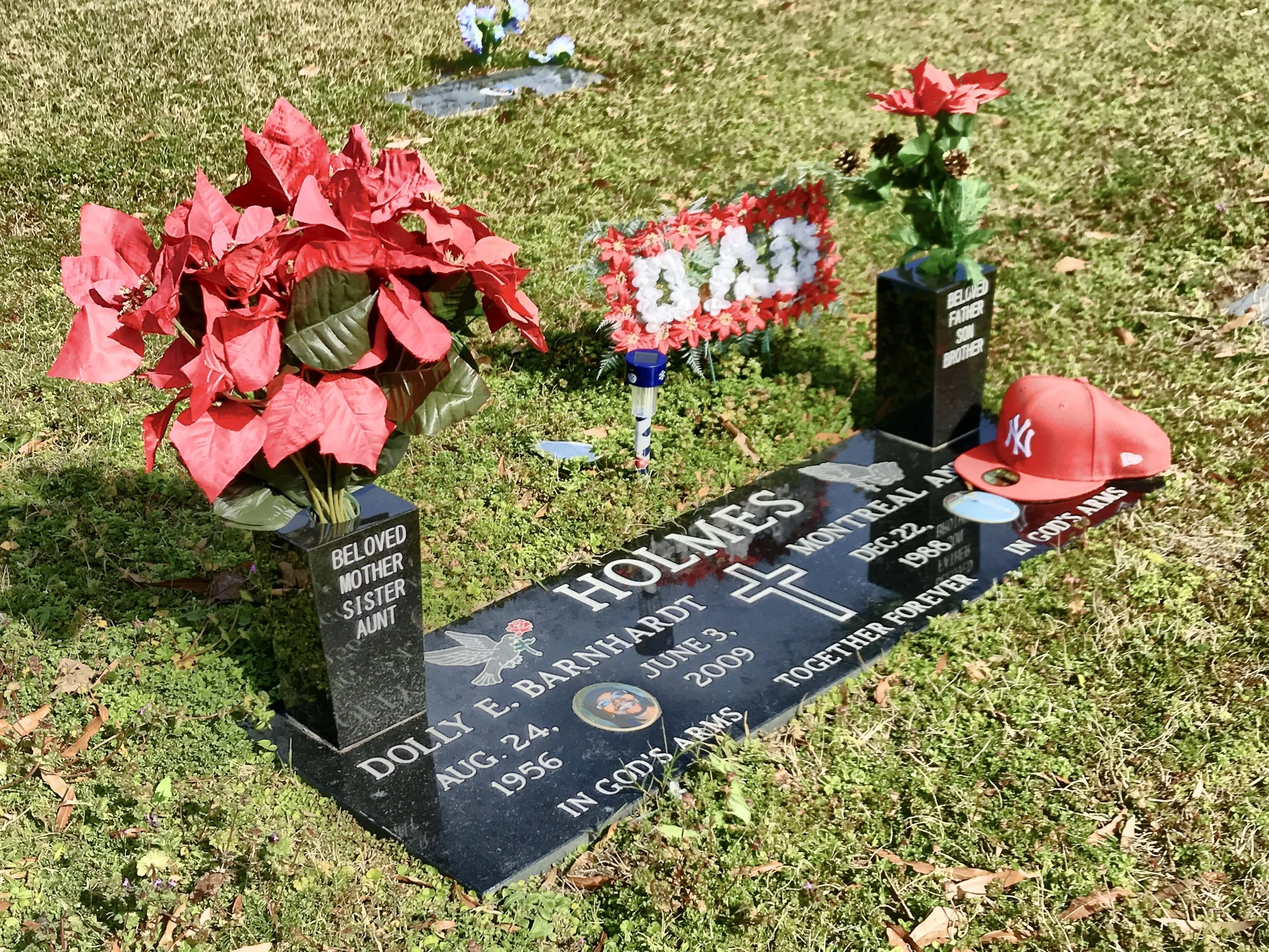 Grave site with red and white floral arrangements, a black headstone reading 'Beloved Mother, Sister, Aunt' for Dolly E. Bartlett, and a red New York Yankees cap placed on the grave.
