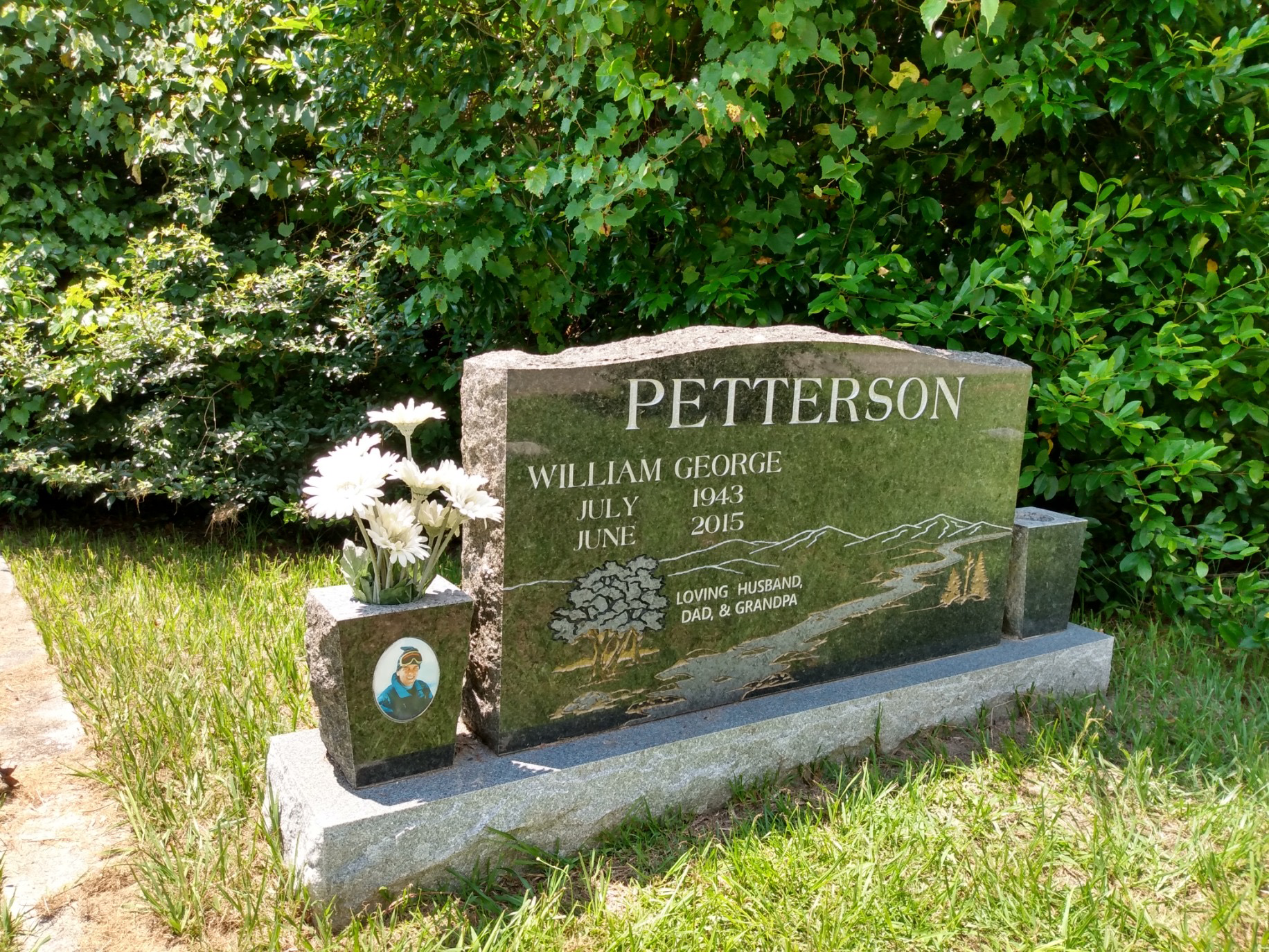 Gravestone for William George Patterson, with flowers and a small photo, surrounded by green bushes and grass.