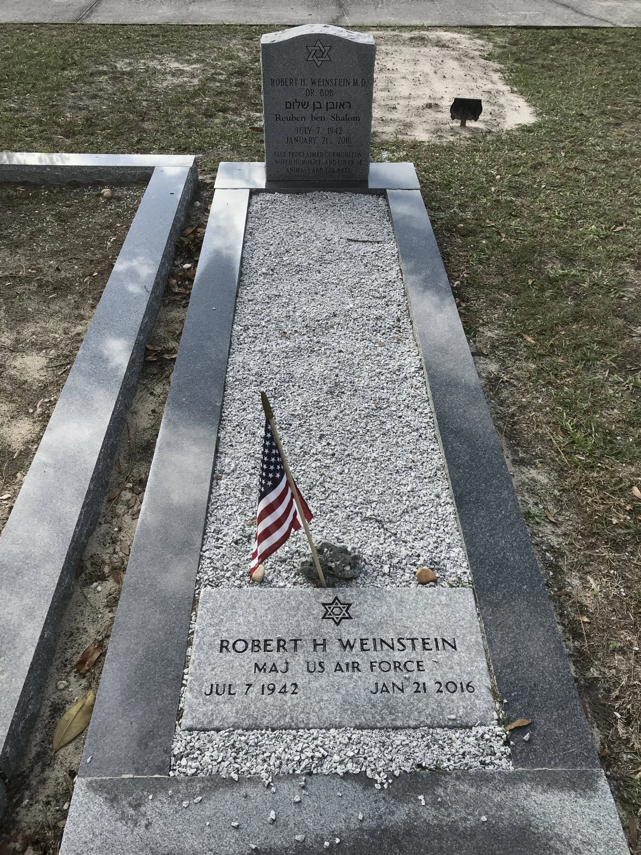 Gravestone for Robert H. Weinstein, M.D., with Jewish symbols. The grave is marked with a white stone with black engraved text. An American flag is placed at the head of the grave. The surrounding area has gravel, dirt, and grass.