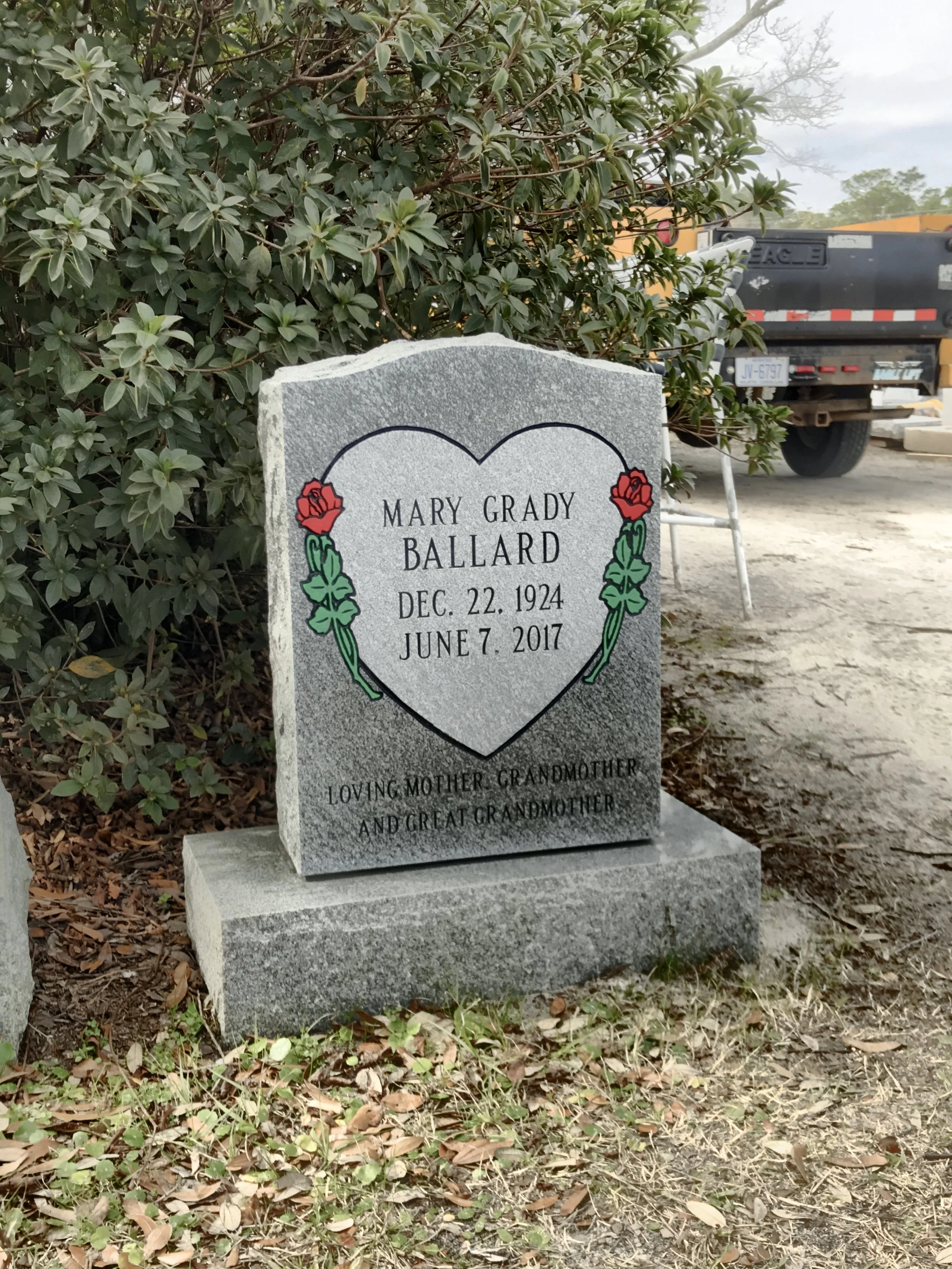 Gravestone with floral decoration in front of bushes and some construction vehicles in the background. The gravestone reads 'Mary Grady Ballard, Dec. 22, 1924 - June 7, 2017,' and mentions loving mother, grandmother, and great-grandmother.