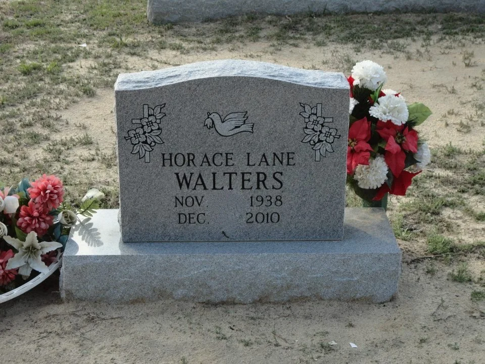 A gray granite headstone with a carved dove and flowers, inscribed with the name Horace Lane Walters, born November 1938 and died December 2010. The headstone is decorated with artificial flowers on each side.