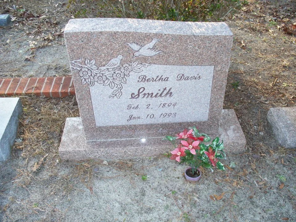 A granite gravestone with an engraved image of two cardsinals and flowers, inscribed with the name Bertha Davi Smith, with birth date February 2, 1894, and death date January 10, 1993, and a small potted pink and green artificial flower at the base.