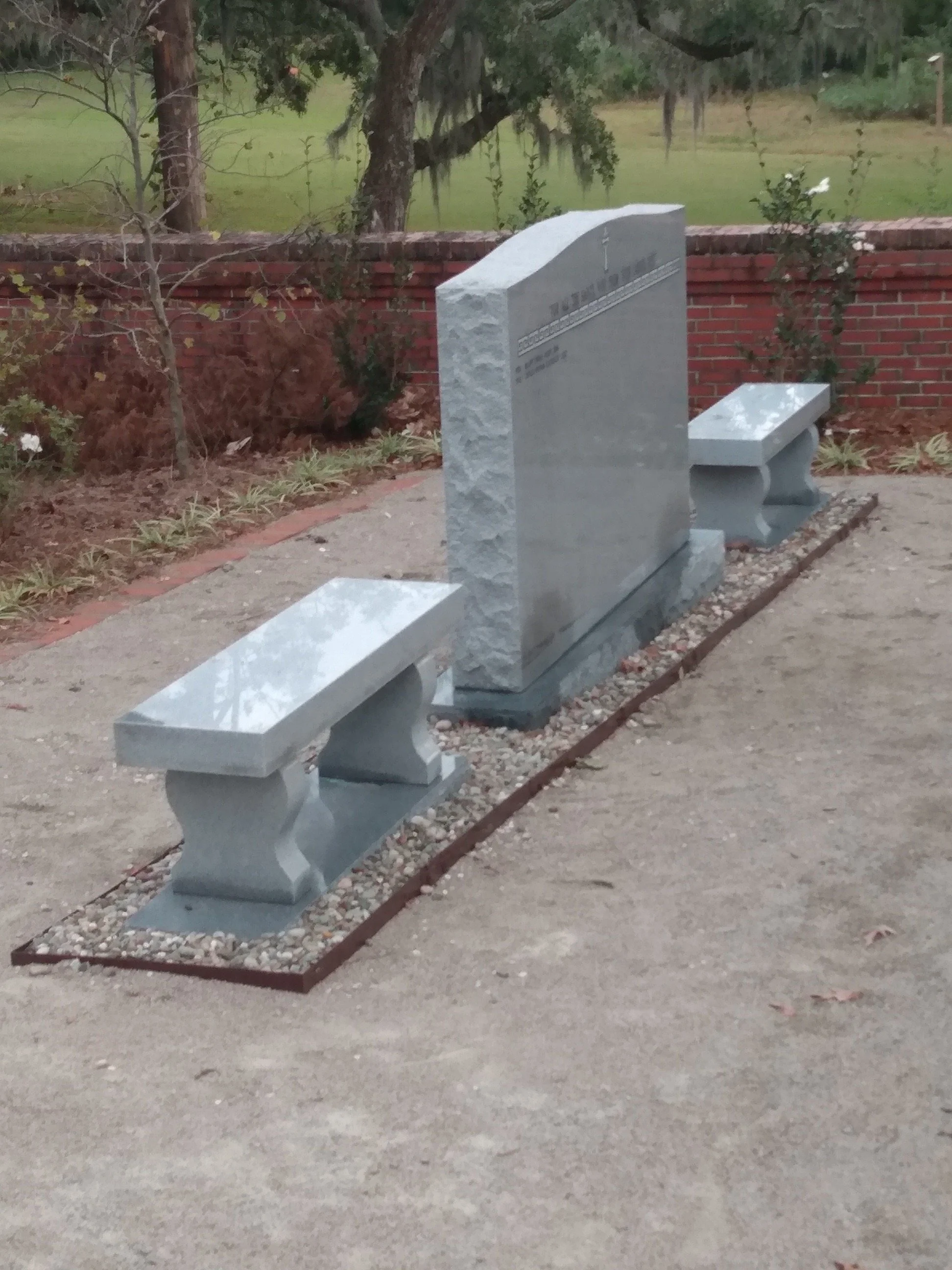 A row of three stone benches with curved bases, placed in front of a brick wall. Behind the wall, there are trees and grassy areas.