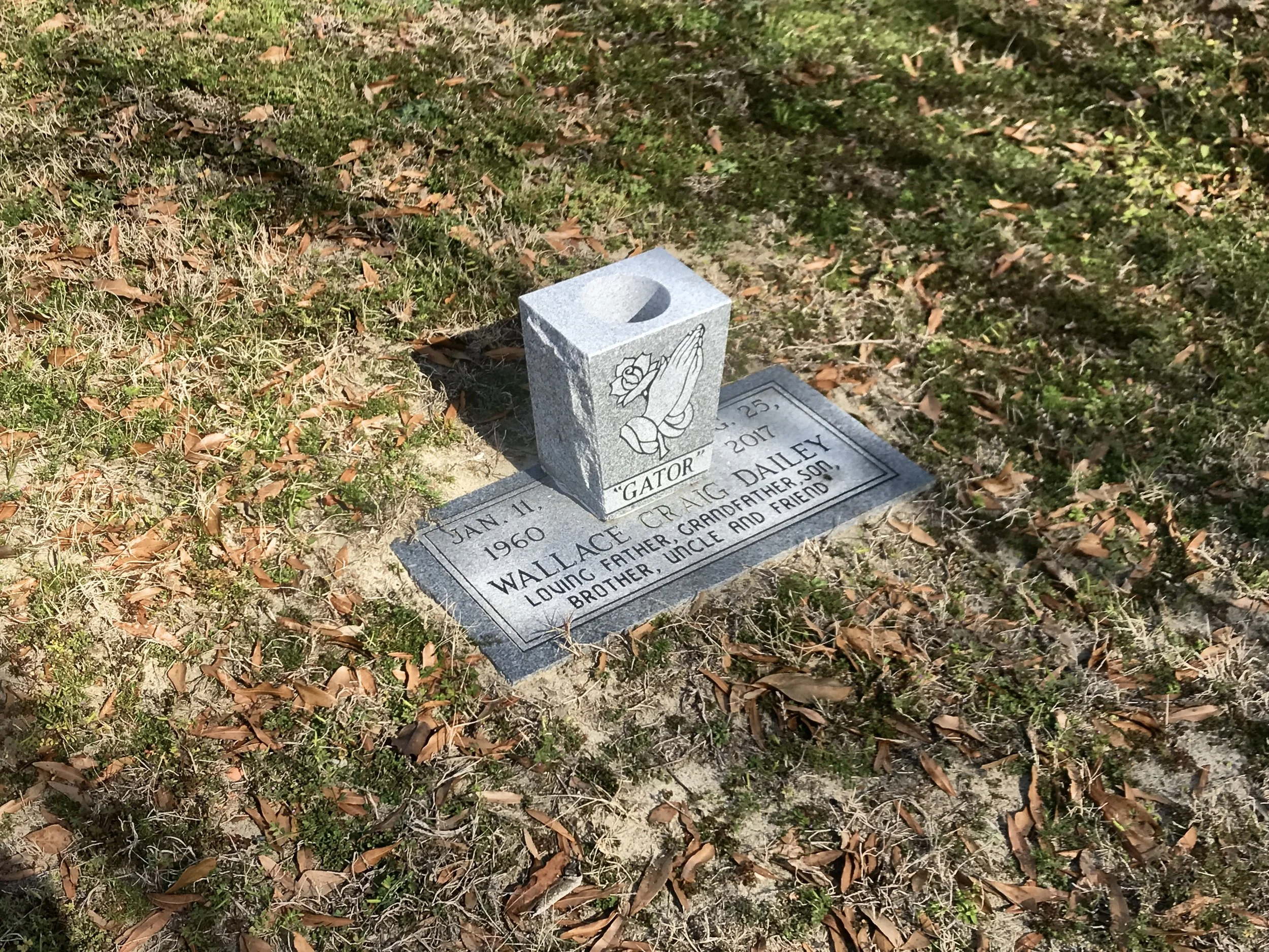 Gravestone with an engraved image of prayer hands and a flower, along with the name Wallace Craig Bailey, a date of January 11, 1960, and a note indicating he is loved by his father, grandfather, uncle, and friends. The gravestone is on the ground wi