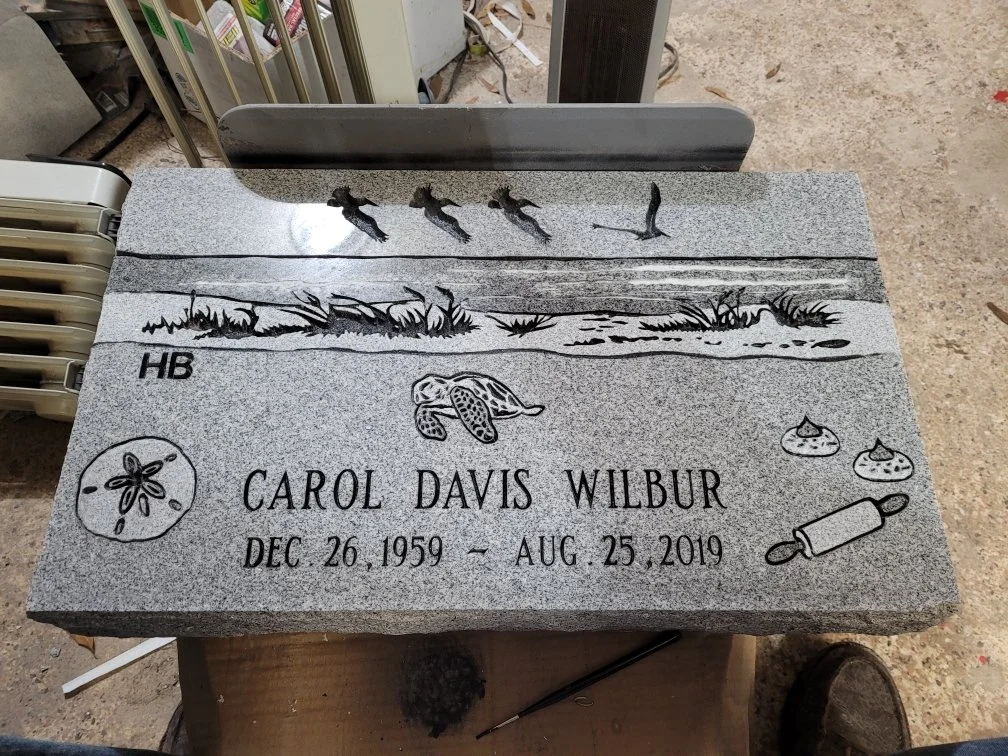 Gravestone for Carol Davis Wilbur, born December 26, 1959, died August 25, 2019, with illustrations of ducks flying, grass, a turtle, cookies, and a rolling pin.