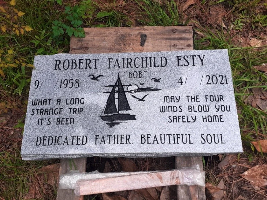 Gravestone with a sailing boat illustration, birds, and sunset or sunrise. It has the name Robert Fairchild Esty, born September 1958, died April 2021. Inscription reads: 'What a long strange trip it's been' and 'May the four winds blow you safely ho