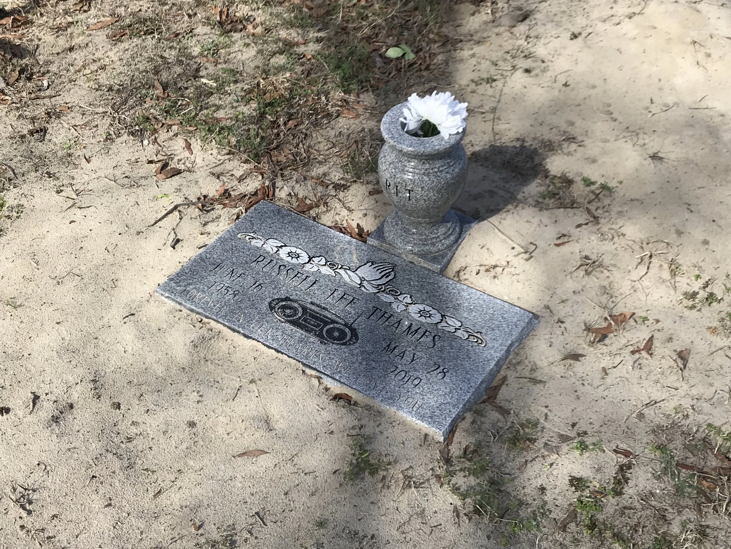 Gravestone with a white flower in a vase next to it, situated in a sandy area with some dry leaves and sparse grass.