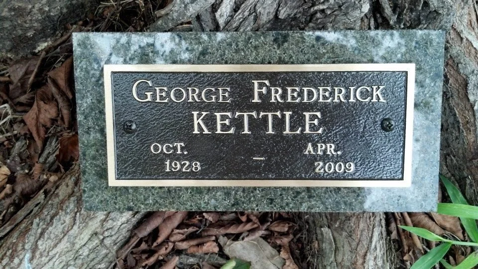 A memorial plaque on a stone base dedicated to George Frederick Kettle, with birth date October 1928 and death date April 2009, mounted at the base of a tree.