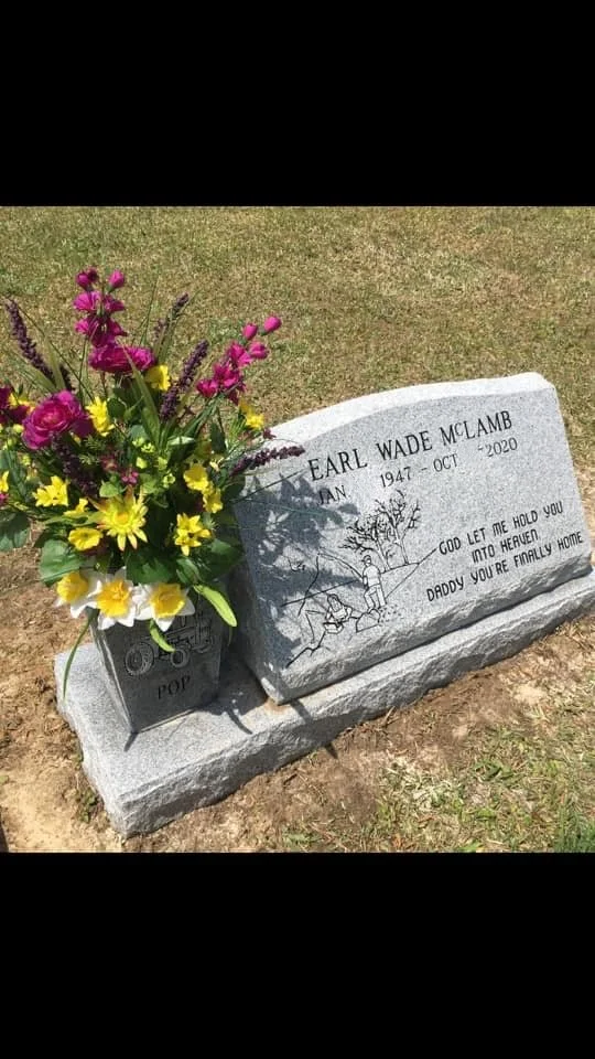 Gravestone with a flower arrangement beside it. The gravestone is for Earl Wade McLamb, who lived from 1947 to October 2020. It has an etched drawing of a family under a tree and an inscription that reads, "God let me hold you into heaven. Daddy you'