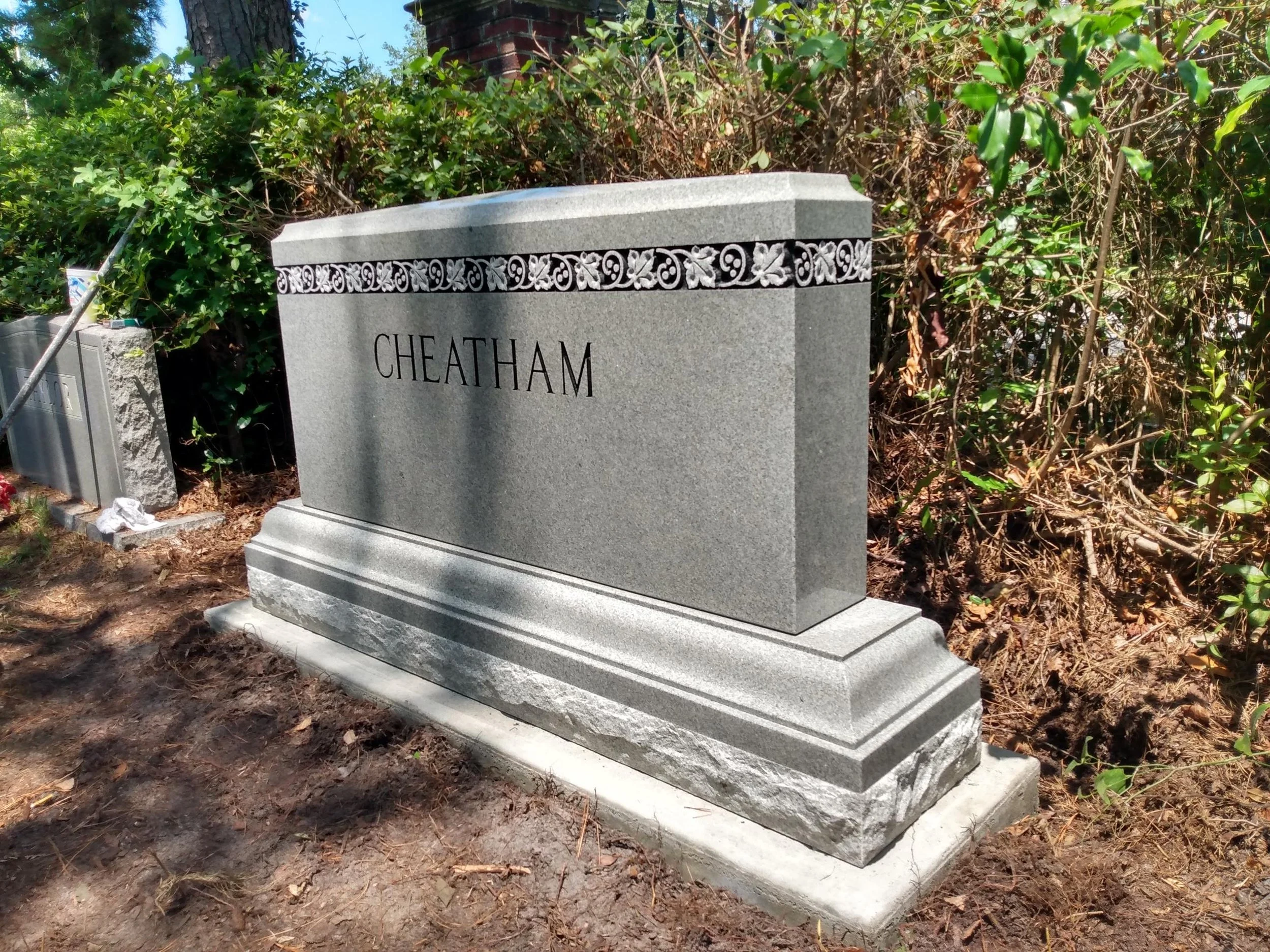 A granite grave marker with the name 'Cheatham' engraved, surrounded by lush bushes and trees.