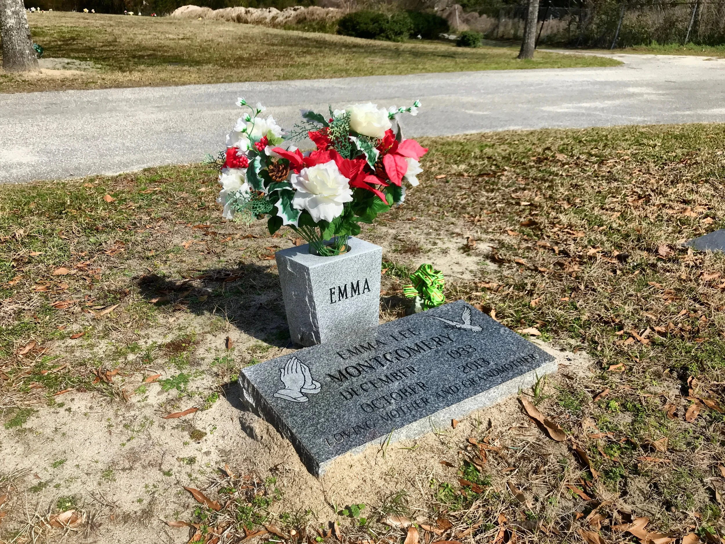 Gravestone at a cemetery for Emma Lee Montgomer, with a flower arrangement consisting of white and red flowers and green leaves, situated on a grassy area.