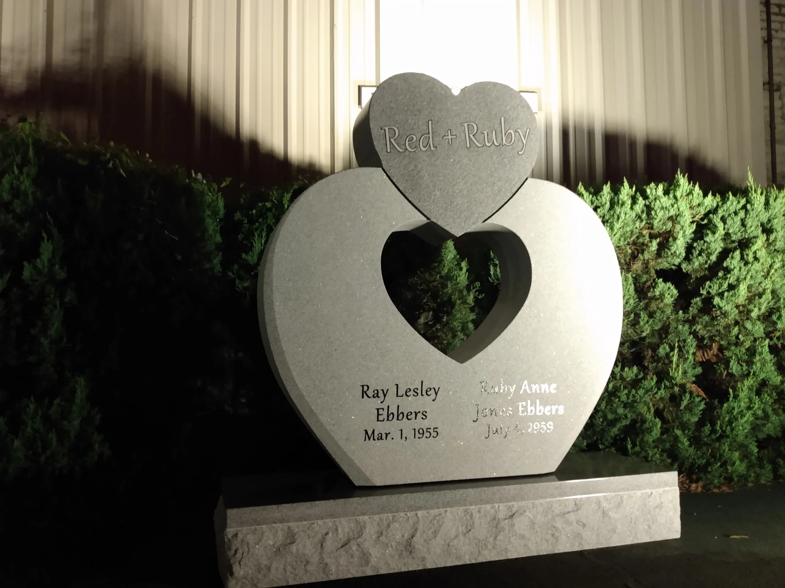 A gray tombstone with a heart-shaped top, engraved with the names 'Red + Ruby', and a smaller white heart-shaped stone in front, engraved with several names and dates, surrounded by green bushes, in front of a beige building at night.