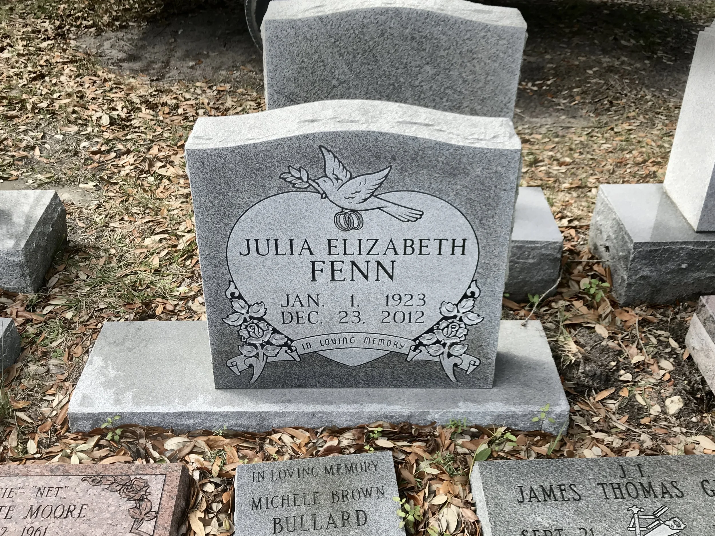 Gravestone for Julia Elizabeth Fenn, born January 1, 1923, died December 23, 2012, with a dove holding an olive branch and wedding rings engraved above her name, located outdoors among other headstones and fallen leaves.