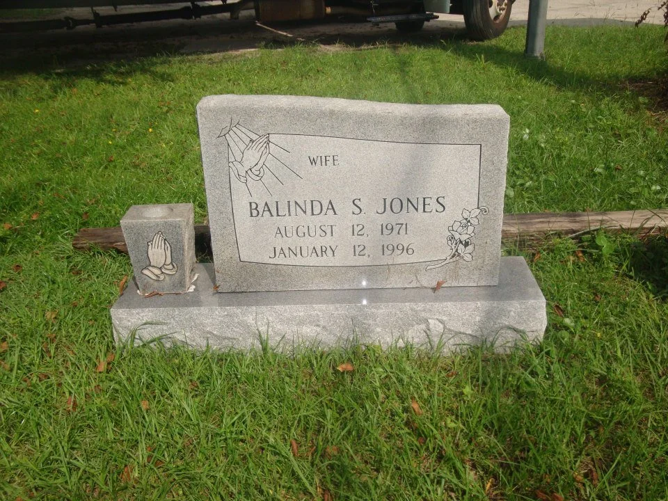 Gray granite tombstone with engraved praying hands and flower, dedicated to wife Balinda S. Jones, born August 12, 1971, died January 12, 1996, situated on a grassy area with a wooden log and a vehicle in the background.