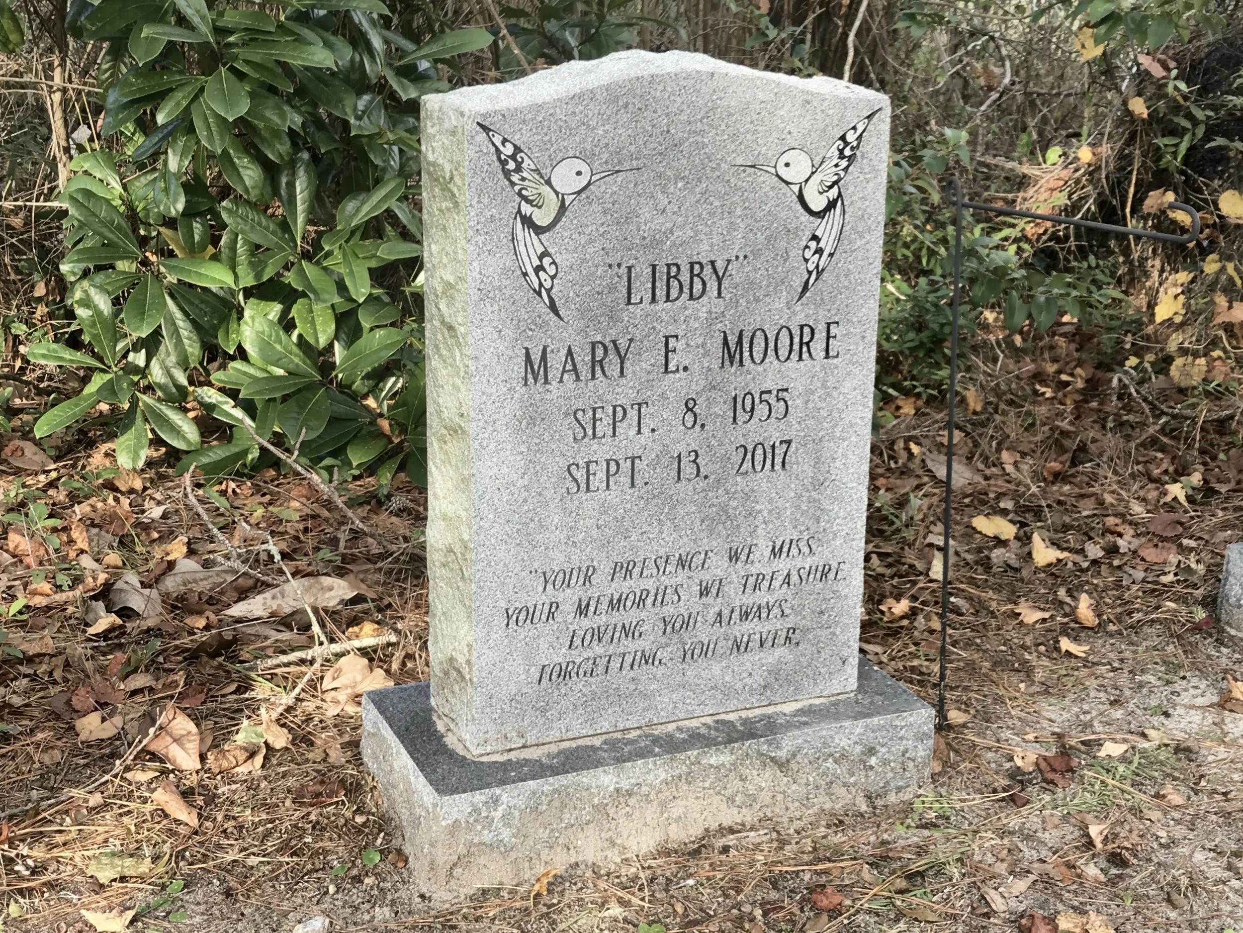 A granite headstone with engraved text and two hummingbird illustrations, surrounded by green leaves, brown foliage, and a black metal fence.