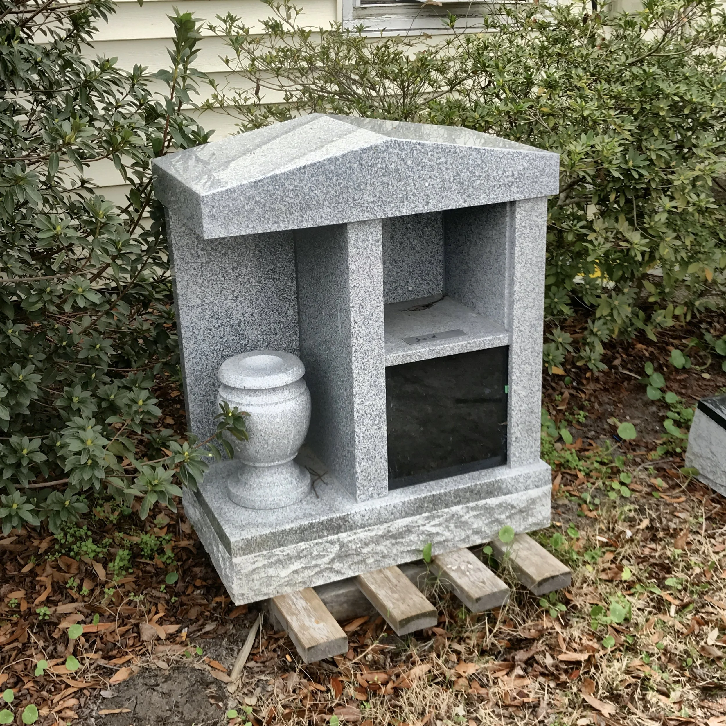 A small granite monument or structure with a flat top and open front, placed outdoors among bushes with green foliage and fallen leaves on the ground.