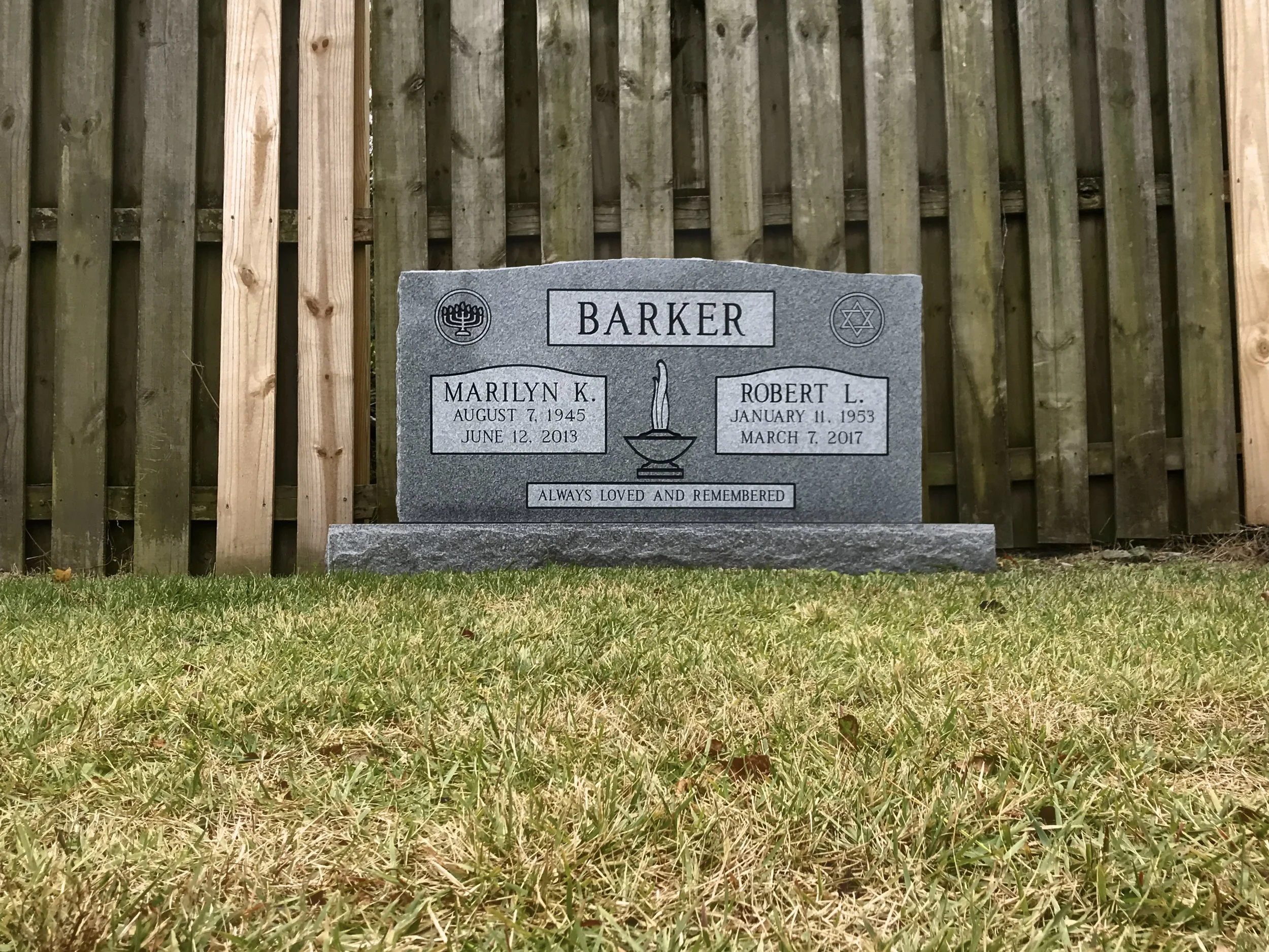 Gravestone with the name Barker, candles, and memorial messages, situated on a grassy area with a wooden fence in the background.