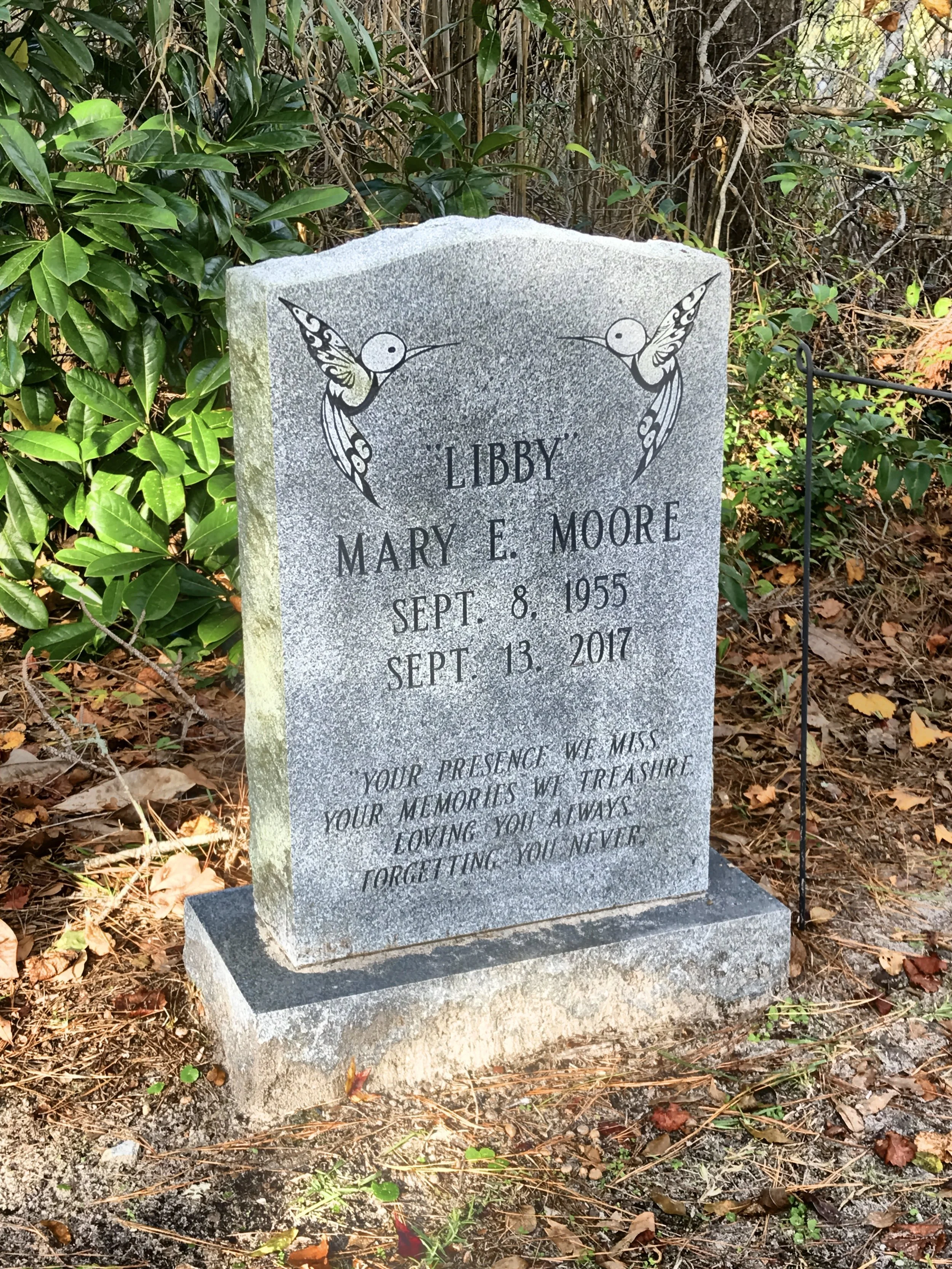 Gravestone with two painted hummingbirds on each side, inscribed with the names Libby and Mary E. Moore, along with birth and death dates, surrounded by green bushes and fallen leaves.