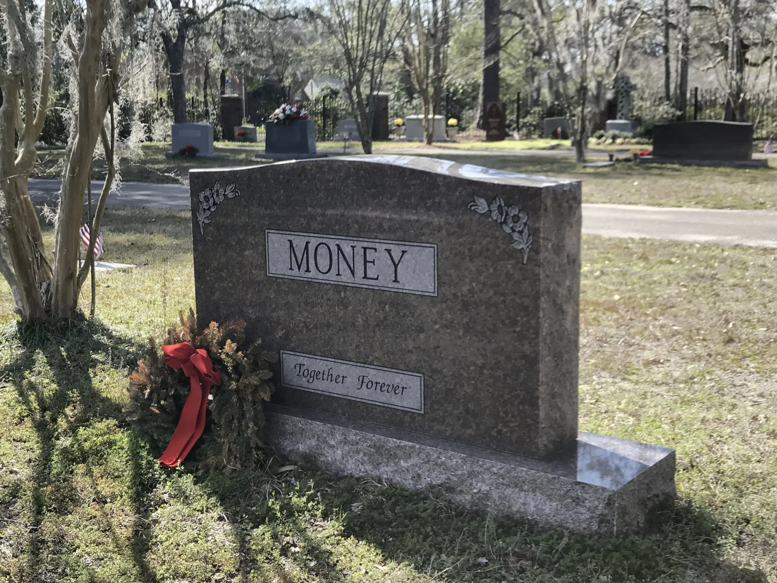 Gravestone in a cemetery with the name 'Money' engraved on it, decorated with floral accents, surrounded by trees and other gravestones, and a small American flag nearby.