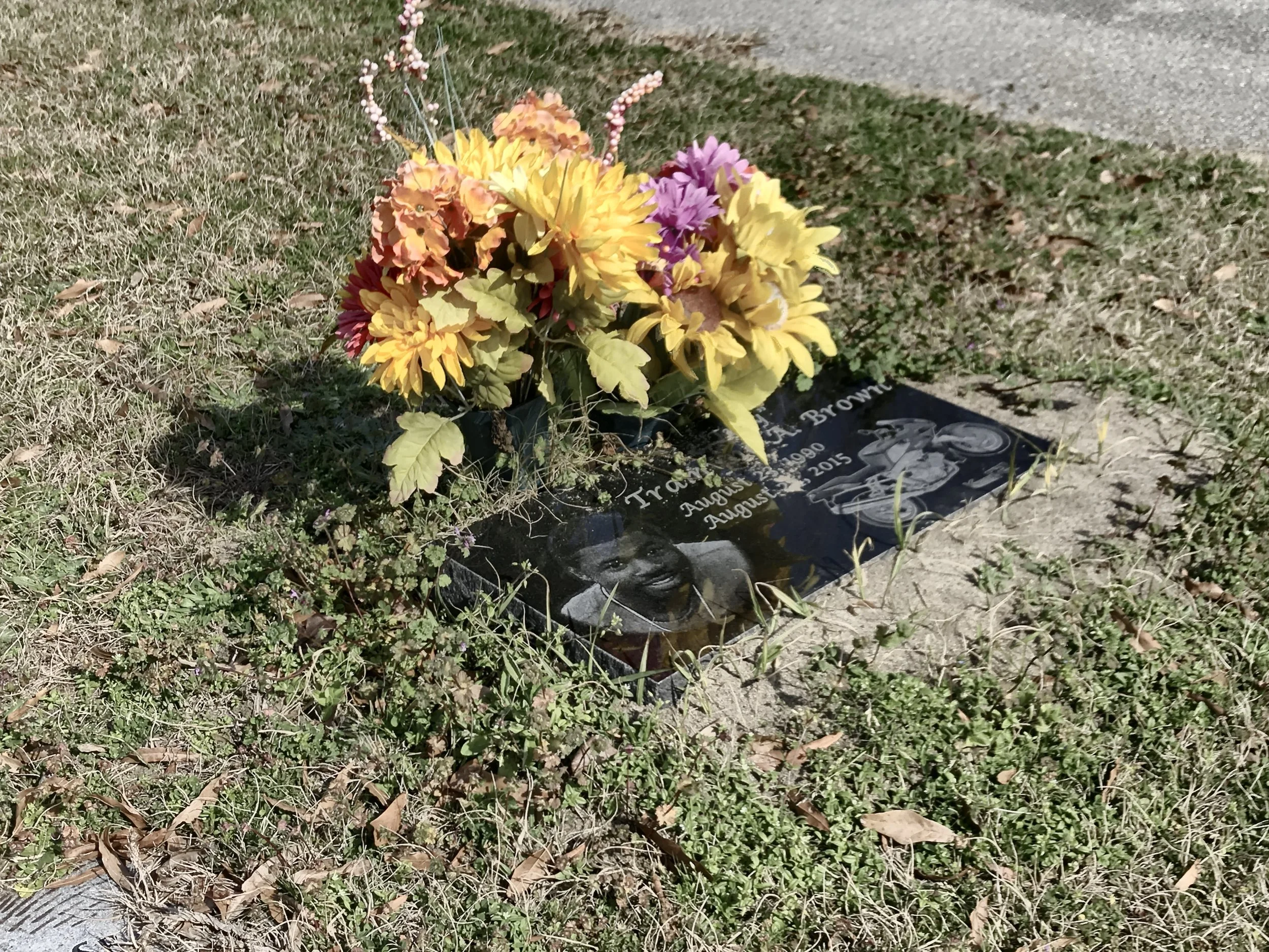 A small bouquet of artificial flowers on a black gravestone in a grassy area with a sidewalk nearby.