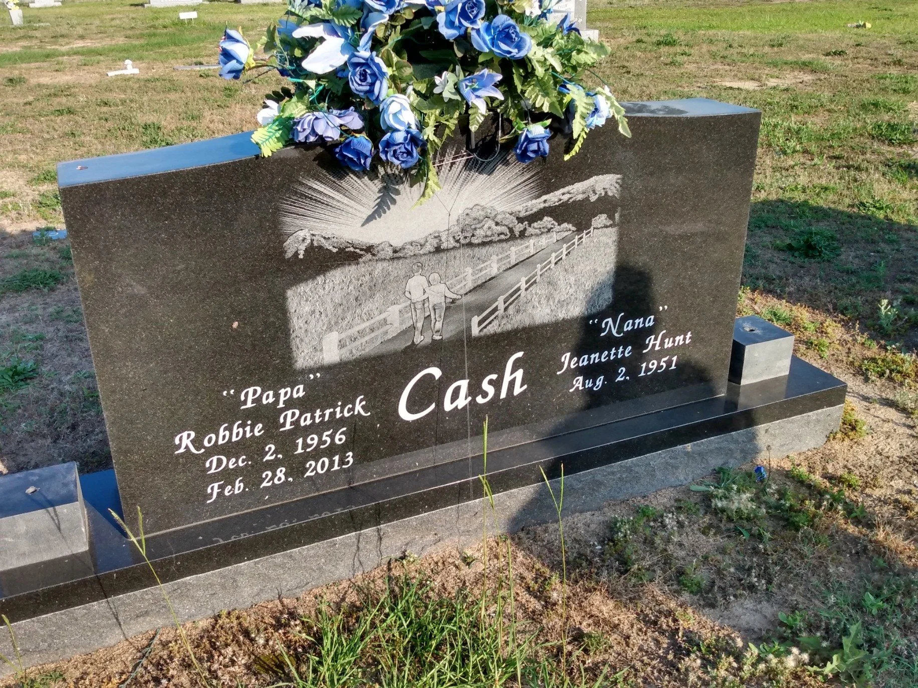 Black granite headstone with engraved image of a couple walking on a bridge with sun rays behind them. The headstone has blue artificial flower decorations on top. It is a grave marker for a person named Cash, with dates of birth and death listed for