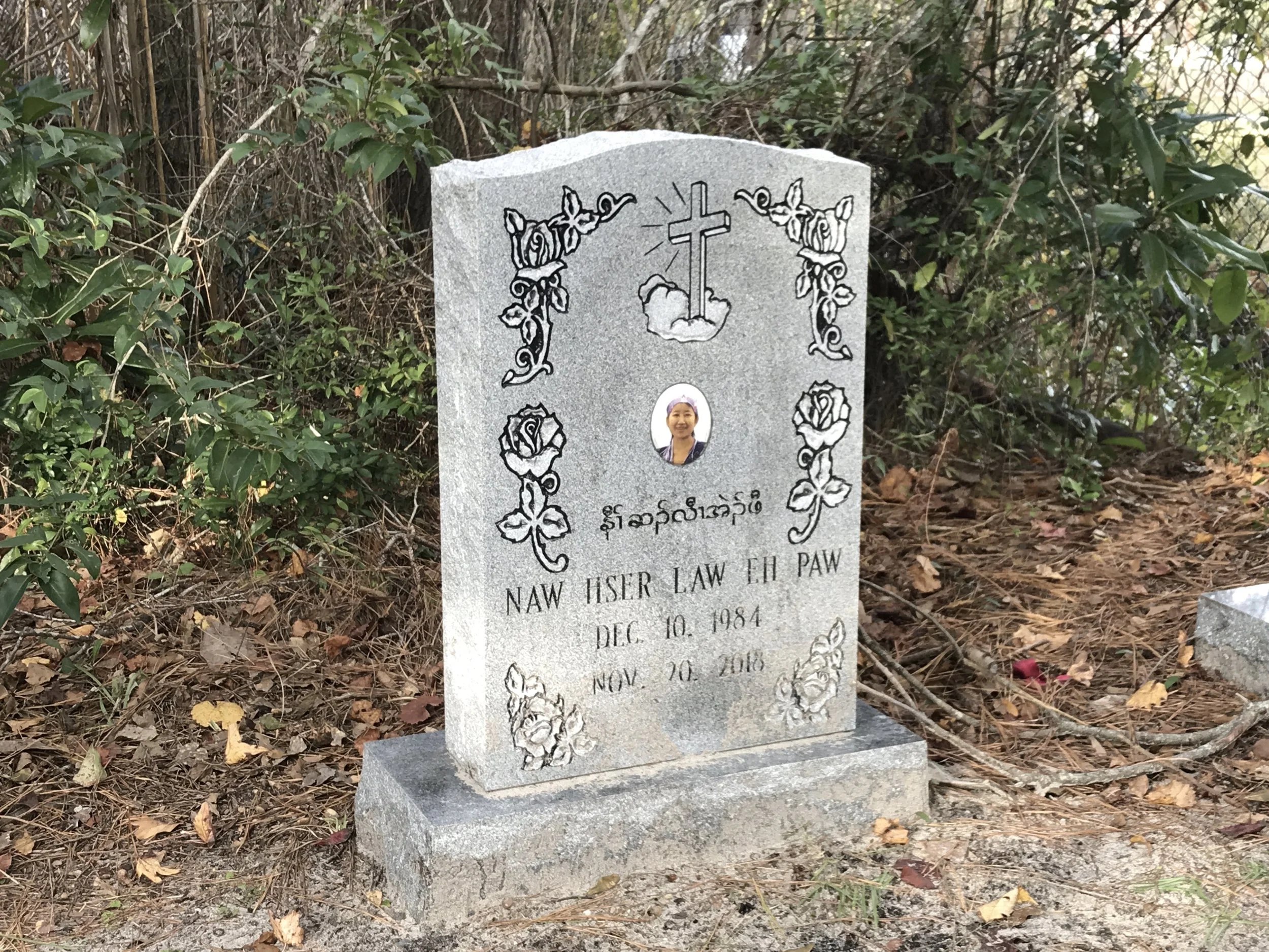 Gravestone with decorative roses and a cross engraving, surrounded by bushes and fallen leaves.